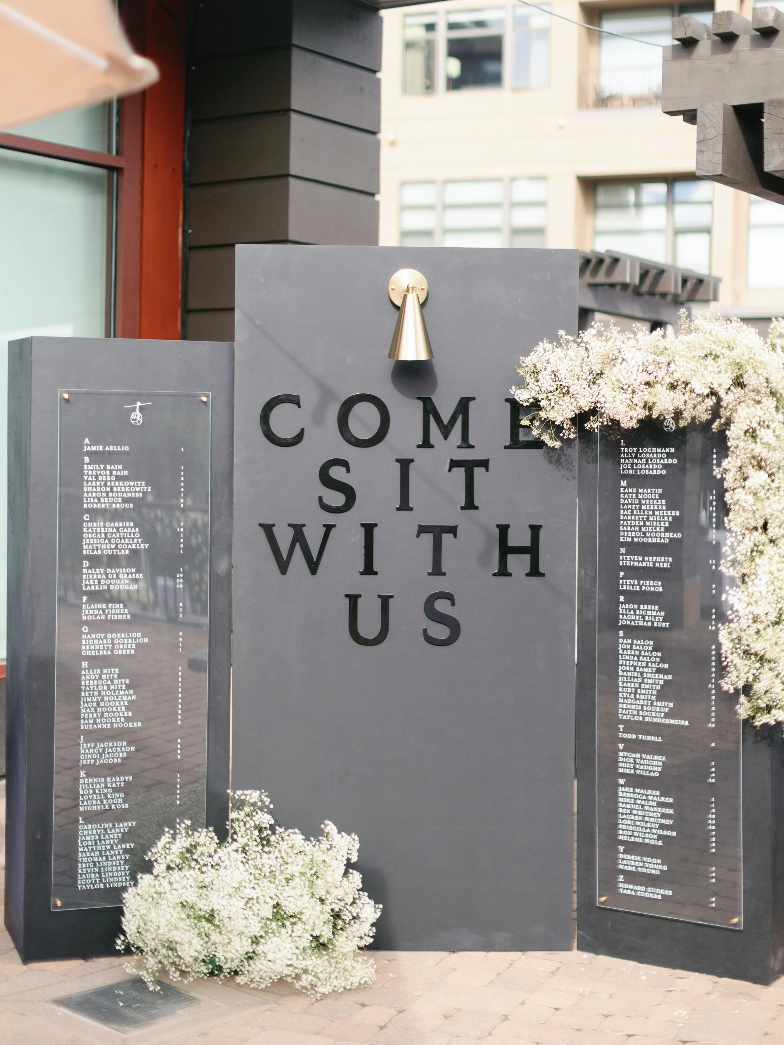 A black outdoor welcome sign with the phrase 'Come Sit With Us' displayed in large black letters, decorated with white flowers on the sides and a gold wall-mounted light above.
