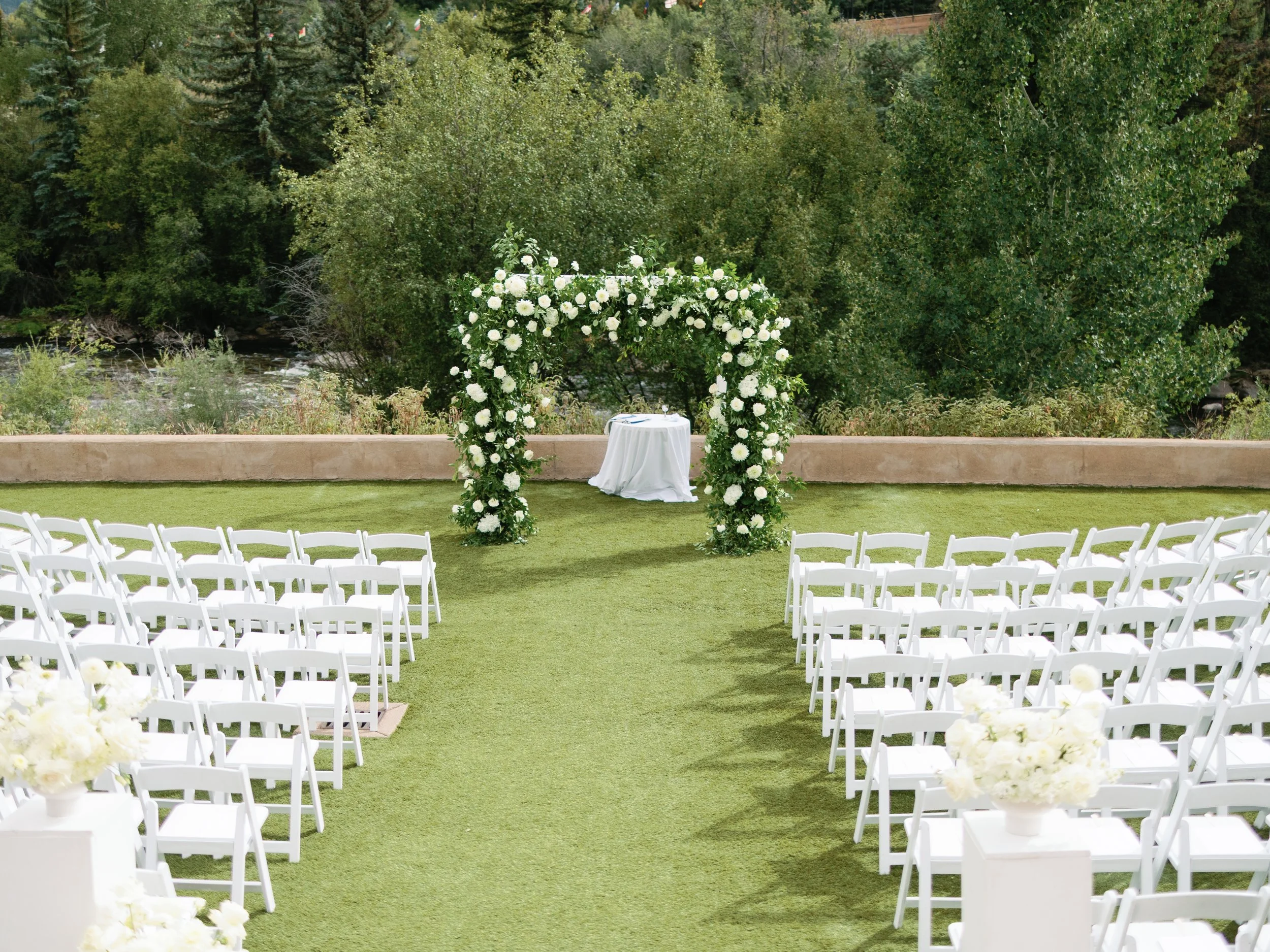 Outdoor wedding ceremony setup with white chairs arranged on green grass, a floral arch at the altar, and trees in the background.