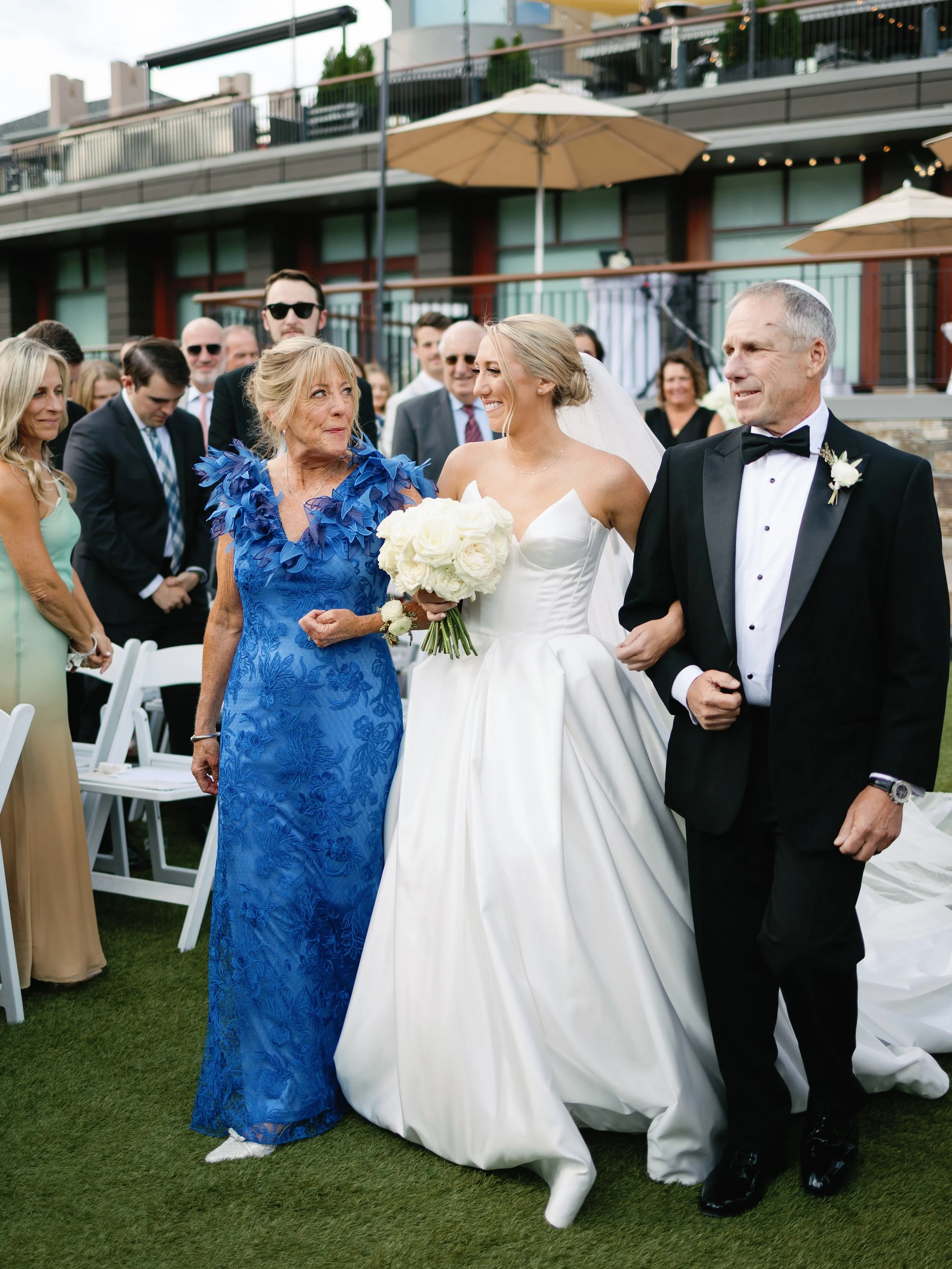 A bride in a white wedding dress holding a bouquet of white roses is walking down the aisle, accompanied by her parents. The mother is wearing a blue lace dress, and the father is in a black tuxedo. Guests are watching and smiling in the background a