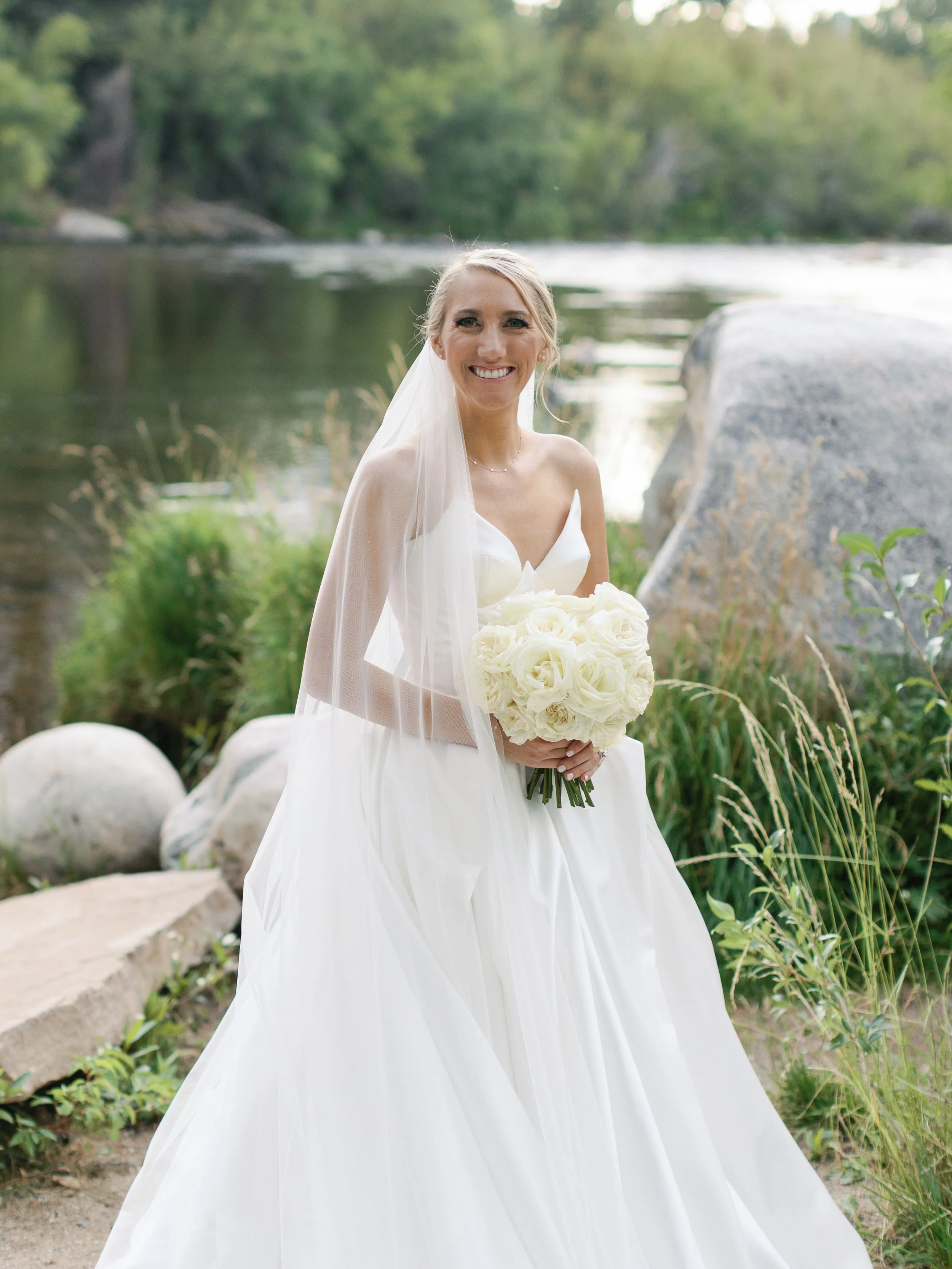 A bride in a white wedding dress holding a bouquet of white roses, standing outdoors near a river with rocks and greenery.