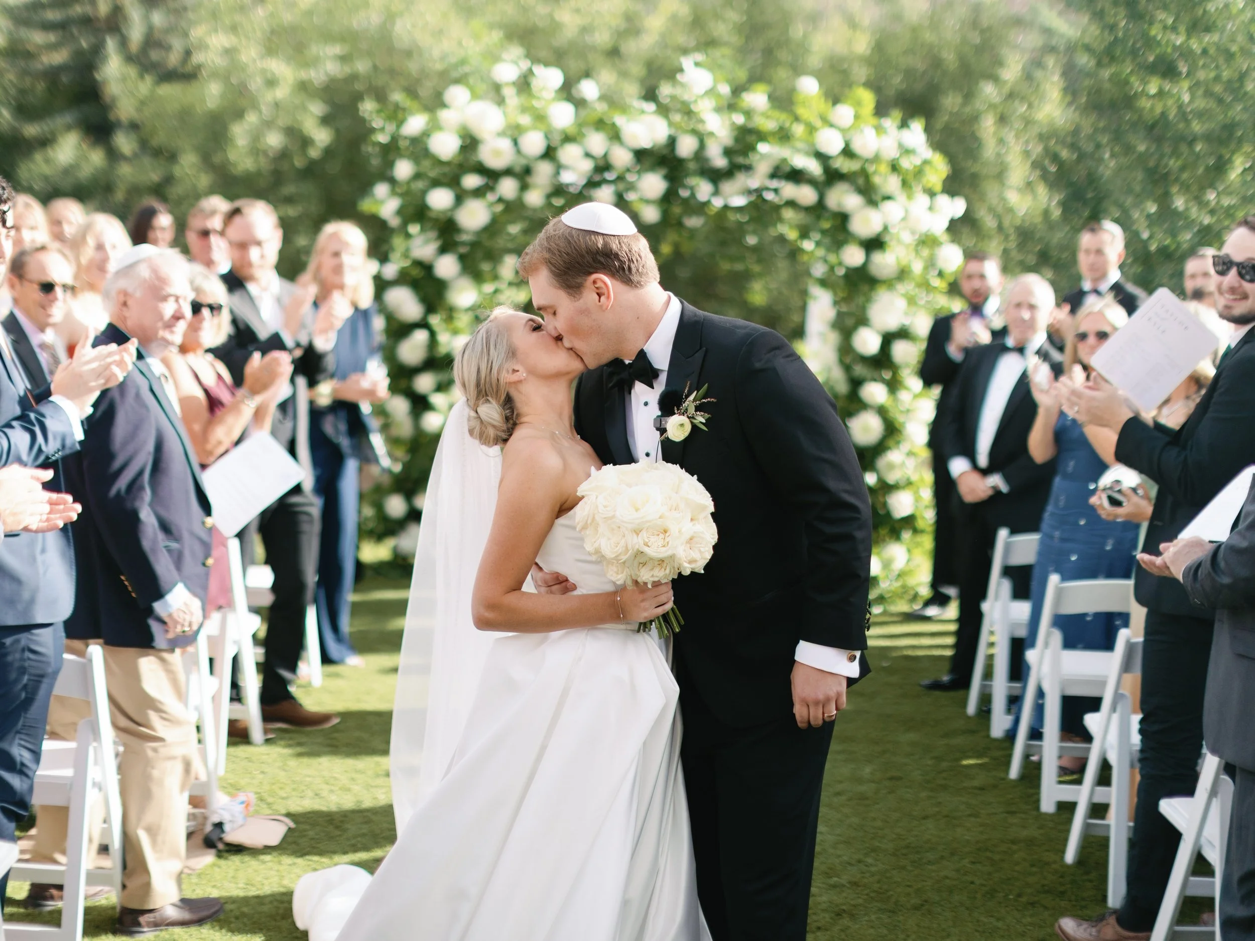 A wedding couple shares a kiss outdoors, with the bride holding a bouquet of white roses, surrounded by guests in formal attire applauding.