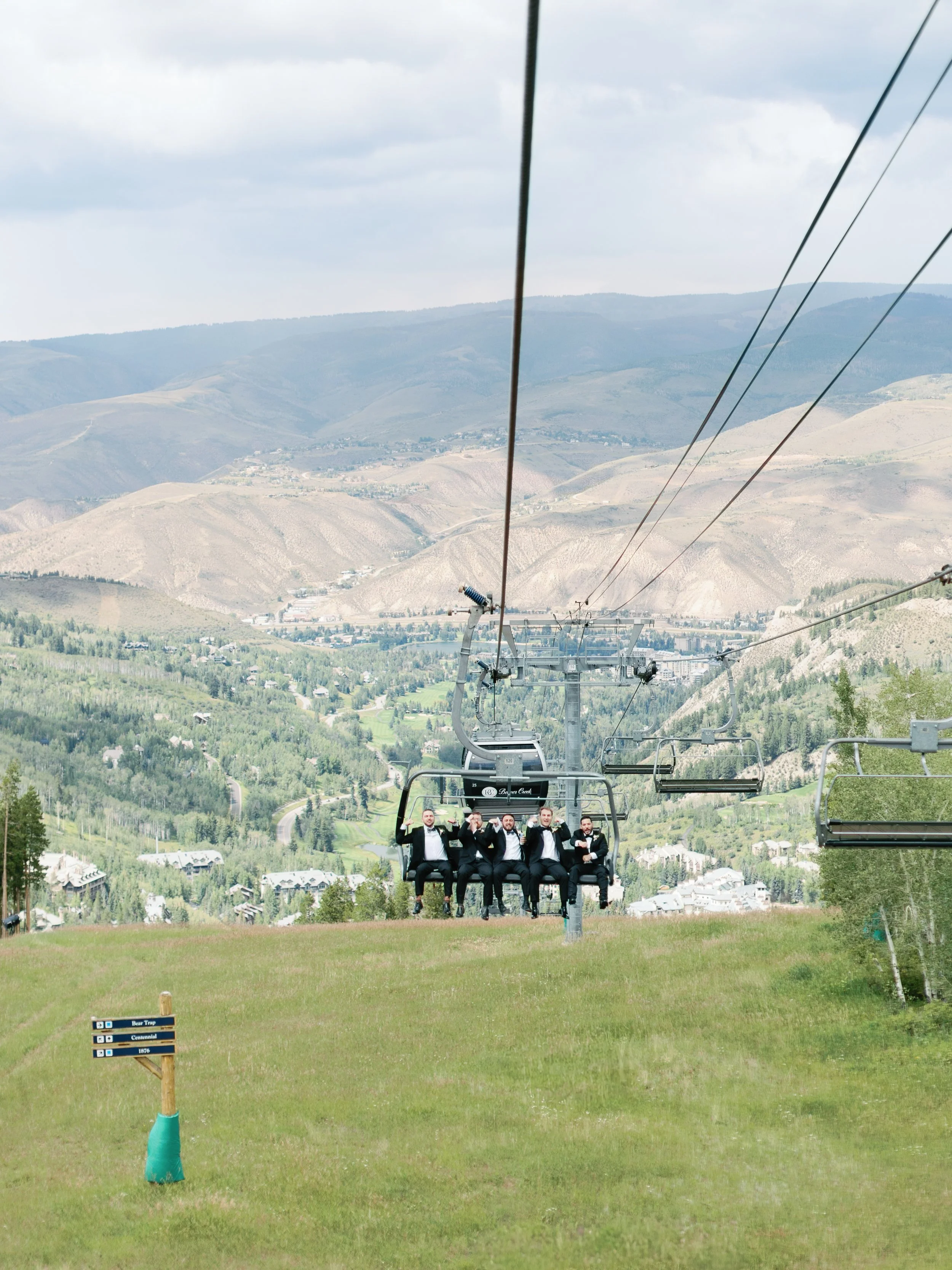Five men in tuxedos riding on a ski lift in a green mountainous area.
