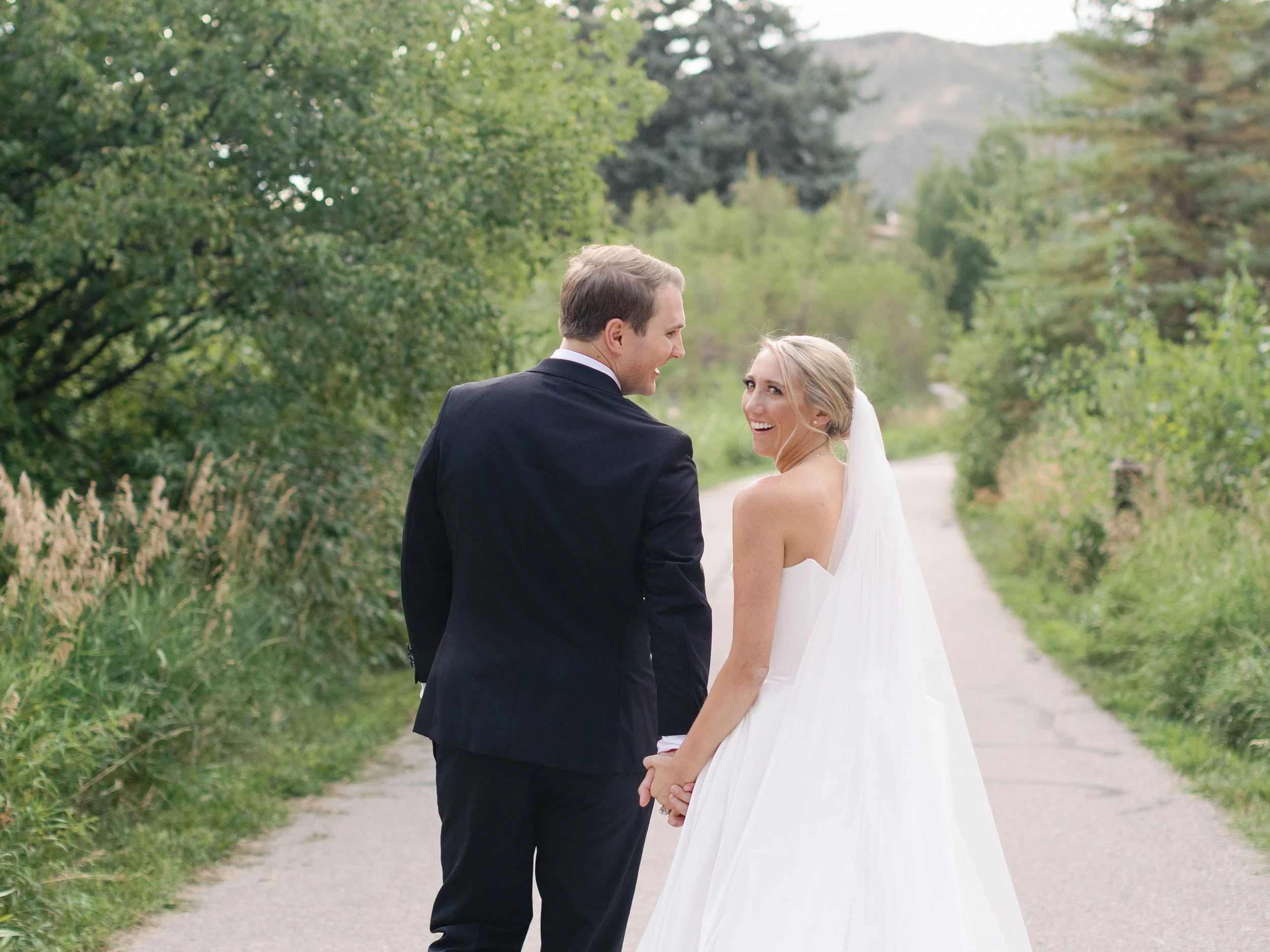 A newlywed couple holding hands and smiling on a country road surrounded by green trees and grass.