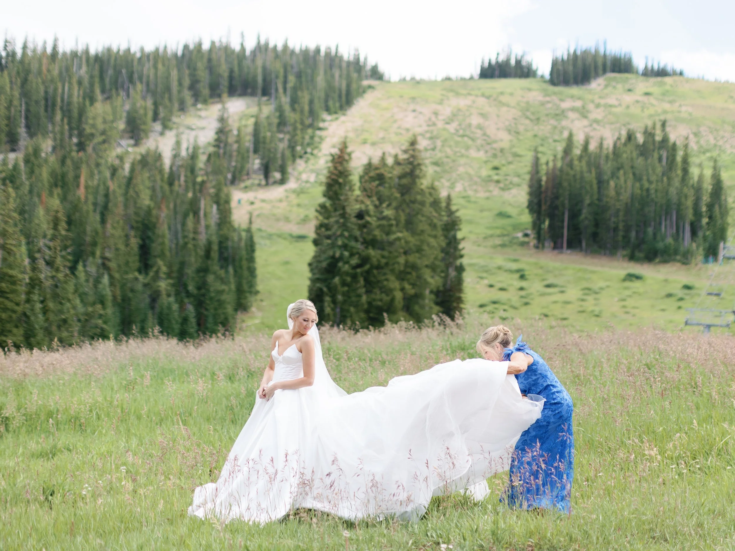 A bride in a white wedding dress standing in a grassy field overlooking a mountain with pine trees, as another woman in a blue dress adjusts her dress.
