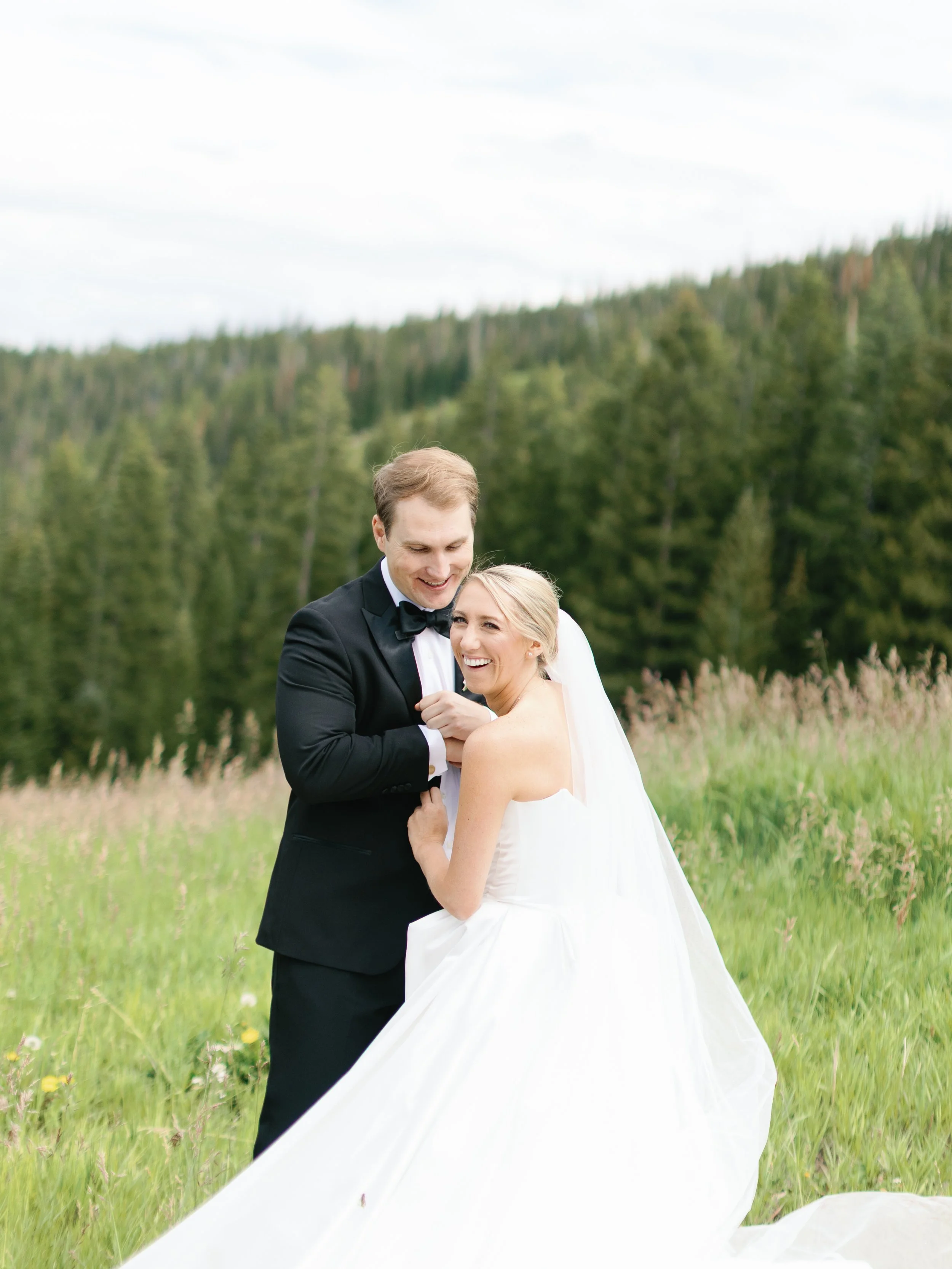 A newlywed couple dressed in wedding attire, smiling and embracing outdoors in a grassy field with a forested hillside in the background.