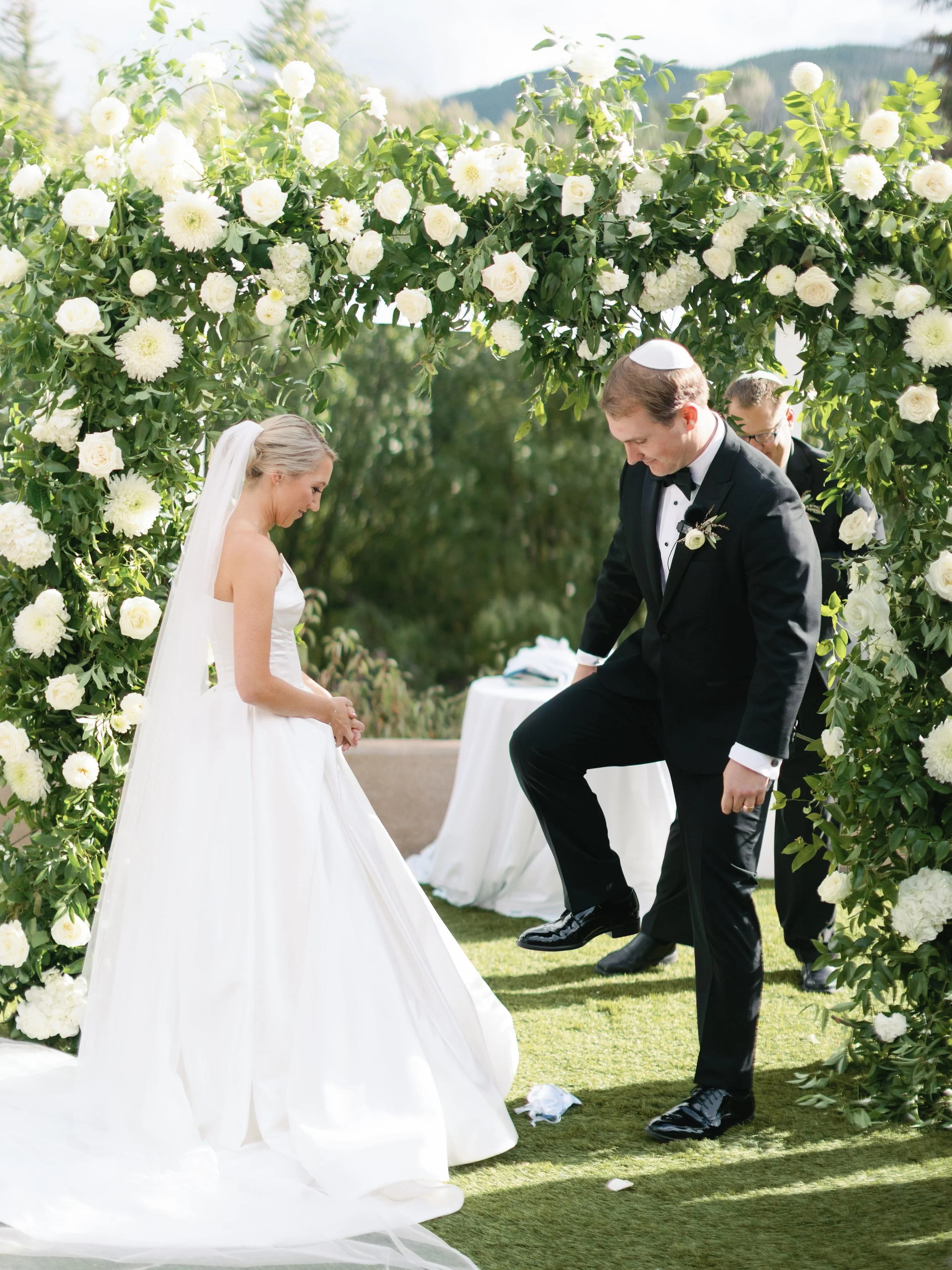 A bride and groom dance under a floral arch at an outdoor wedding ceremony on a sunny day with mountains in the background.