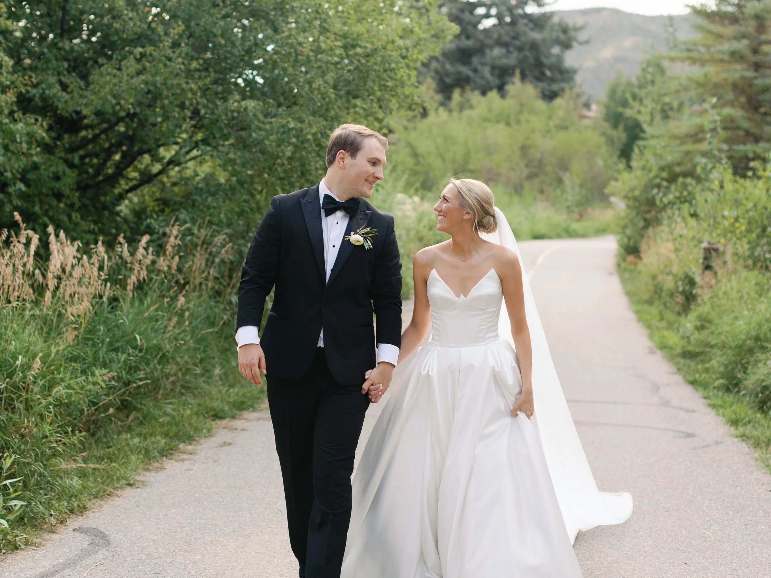 A bride and groom walking hand in hand outdoors on a paved path surrounded by greenery, smiling at each other.