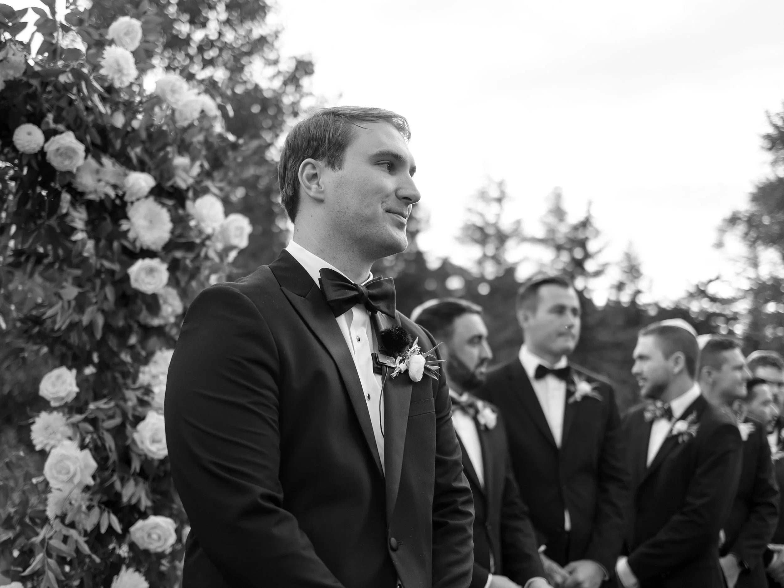 A group of men dressed in black tuxedos with bow ties, standing outdoors at a wedding ceremony. One man in the foreground is smiling, with a boutonniere on his lapel, and the others are standing in the background near floral decorations.