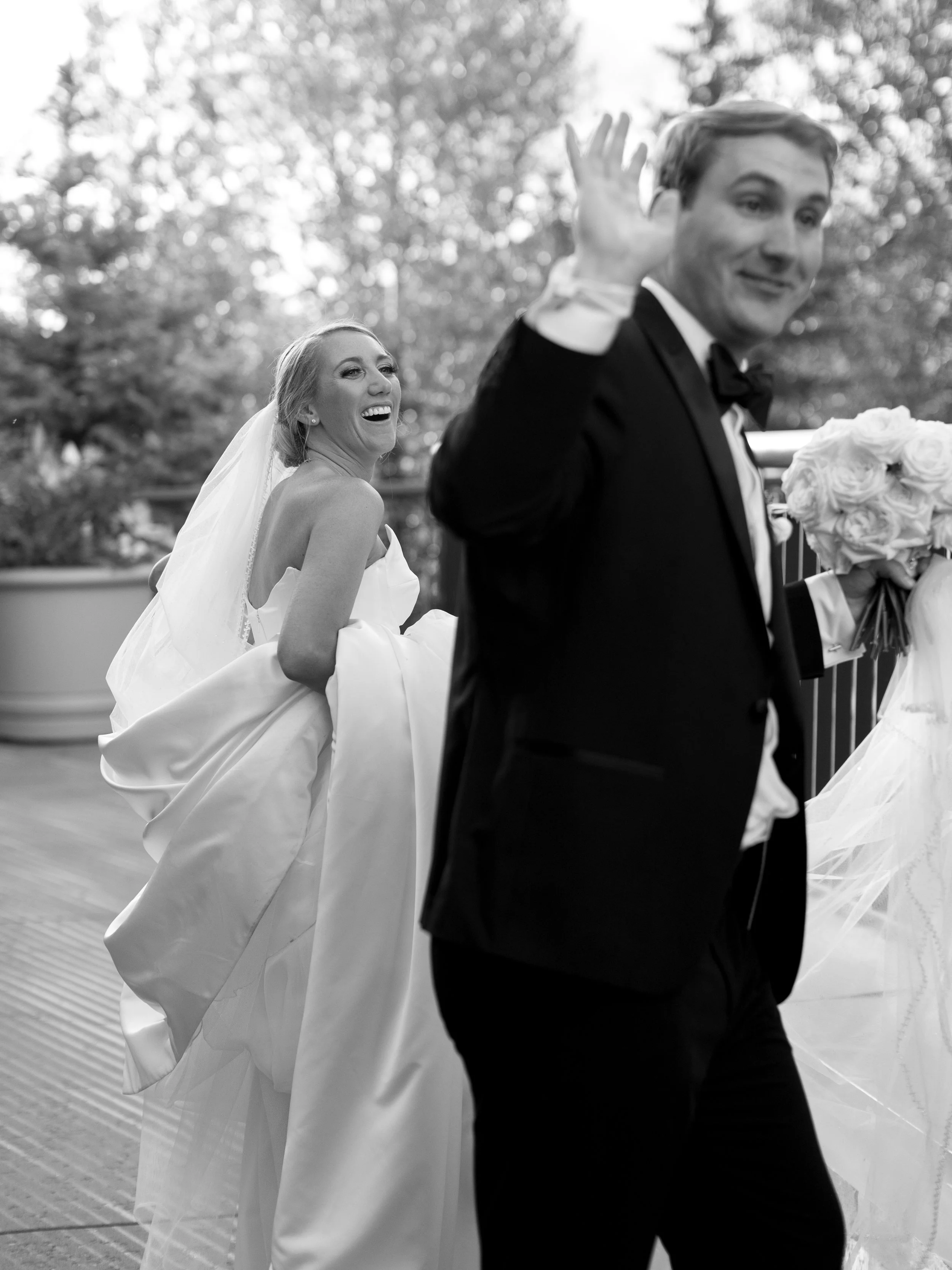 A bride and groom at a wedding, with the bride laughing and the groom waving, as they walk outdoors.