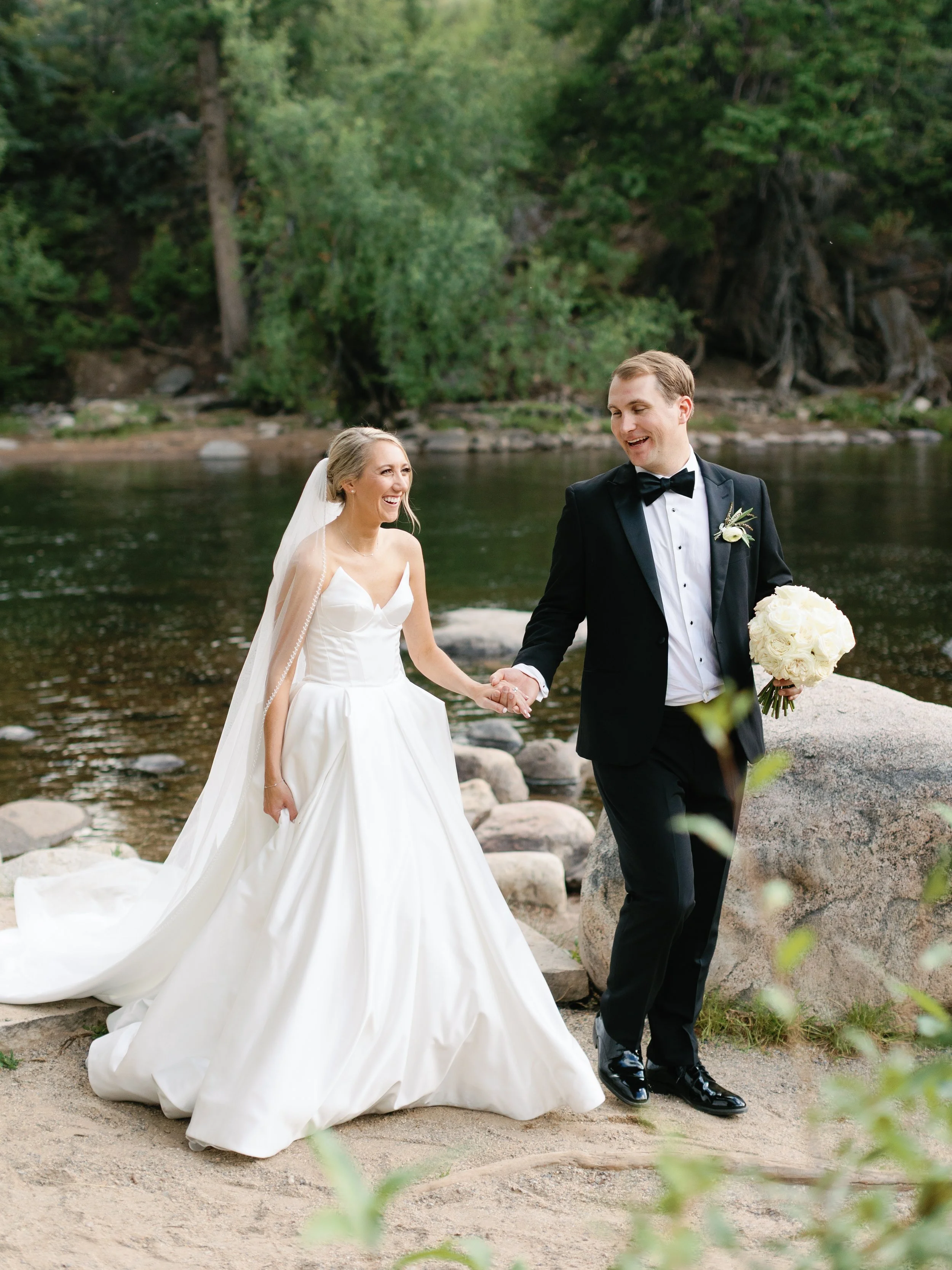 A newlywed couple walking hand in hand by a river, surrounded by trees and rocks, smiling at each other.