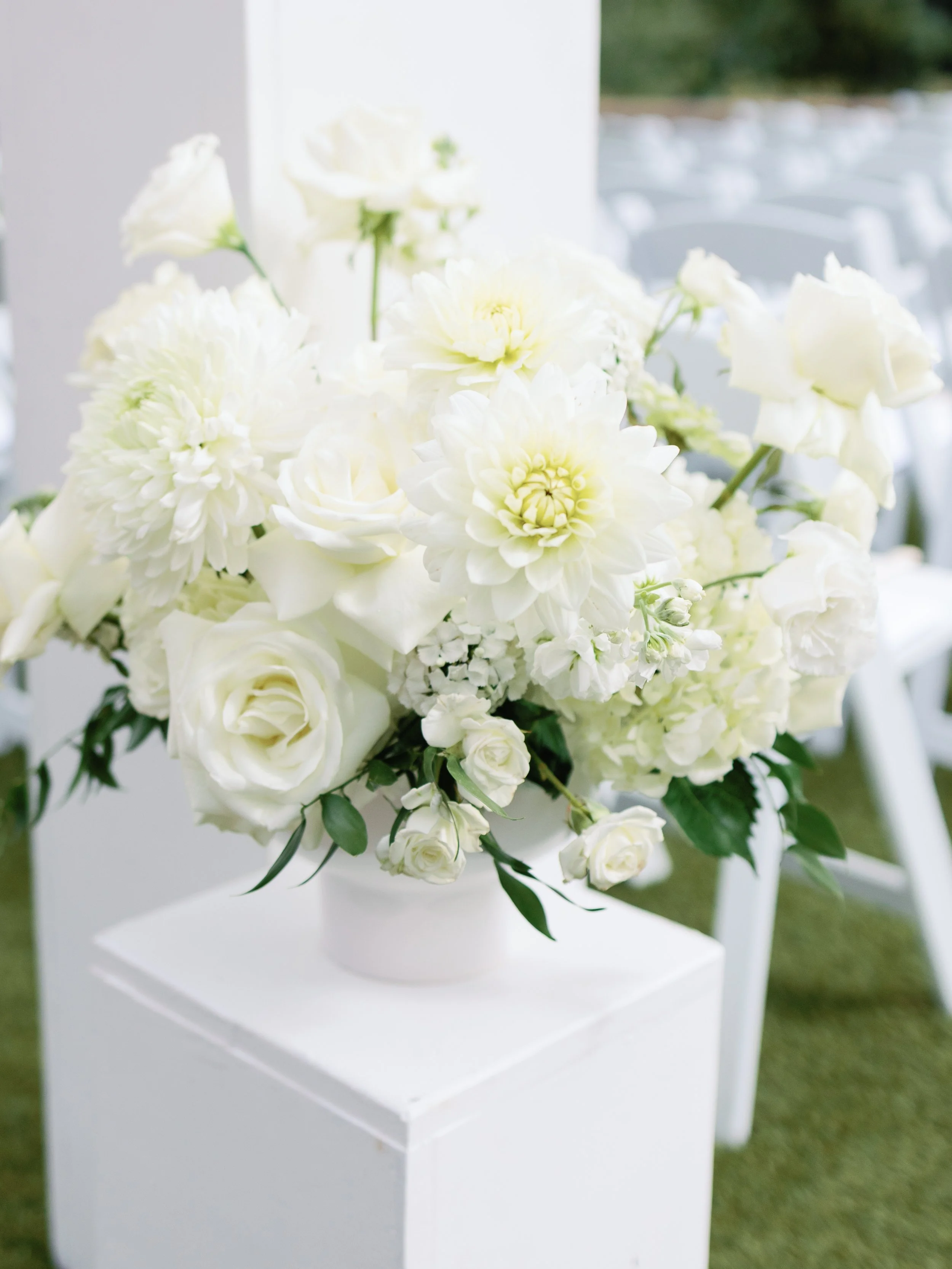 A white flower bouquet composed of roses, dahlias, and other white blooms, displayed in a white vase on a white pedestal.