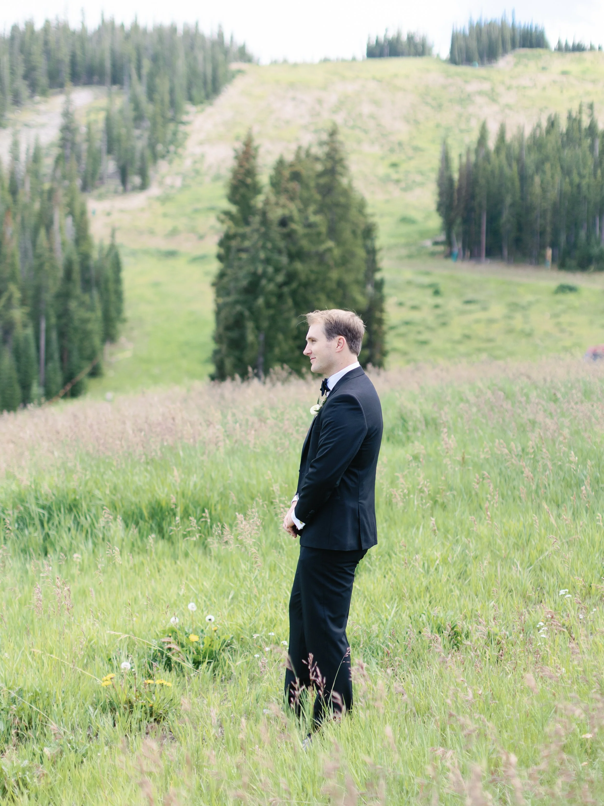 A man dressed in a black tuxedo standing in a grassy field with trees and mountain hills in the background.