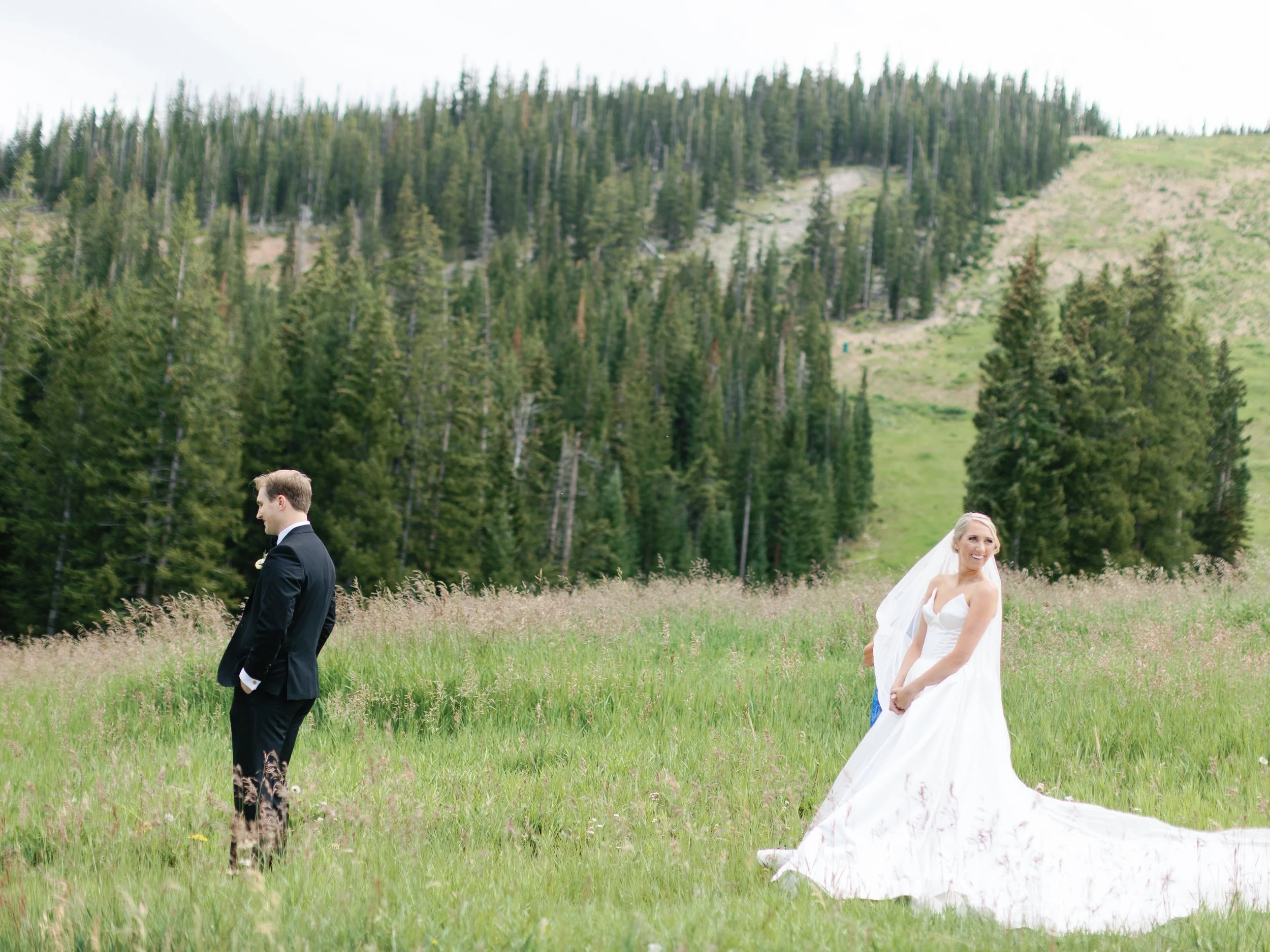 Bride in a white wedding dress and veil smiling with her hands near her waist, while groom in a black suit stands facing away in a green grassy field with a forested mountain in the background.