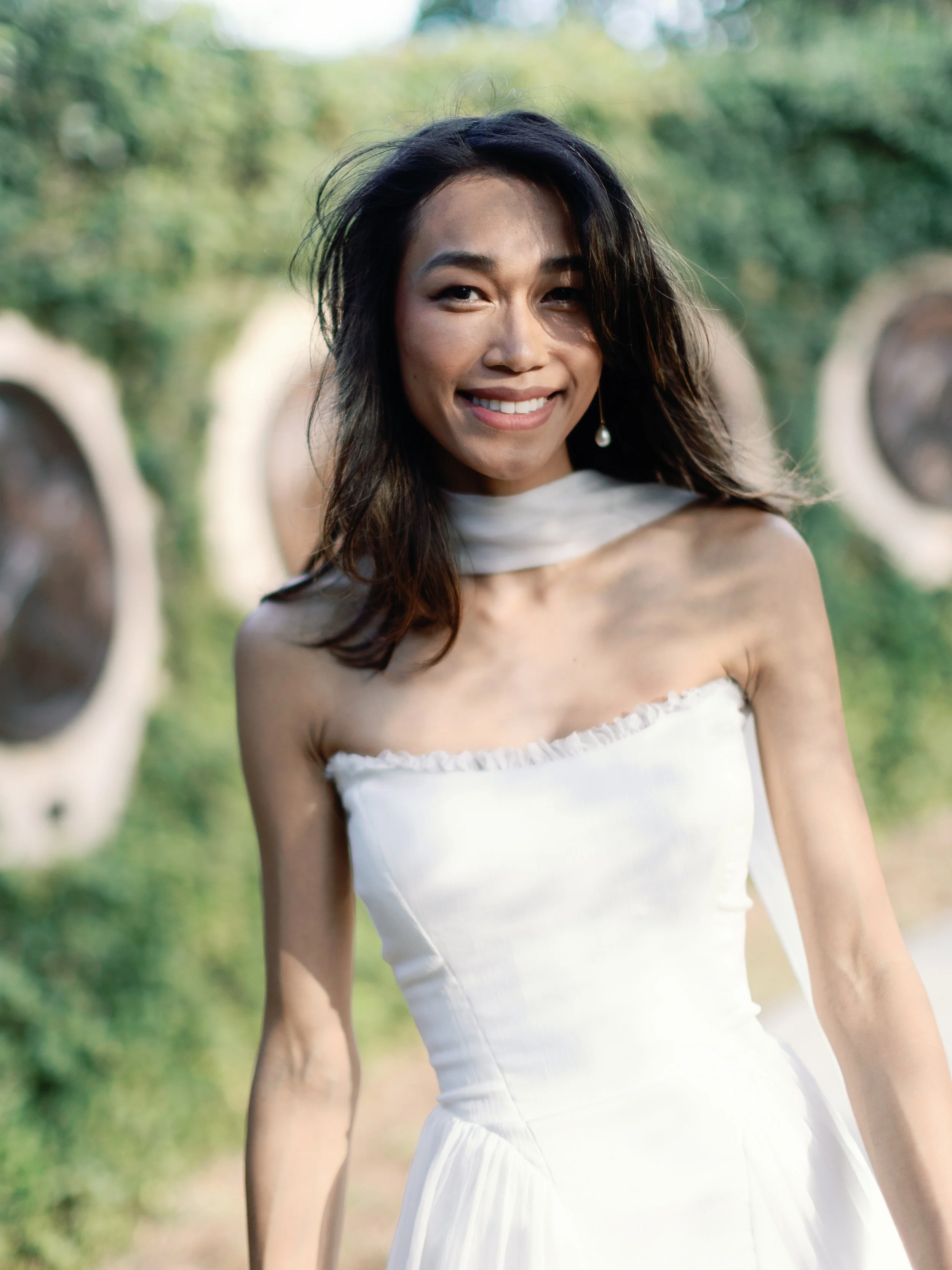 A smiling woman wearing a white strapless dress outdoors with greenery and large circular structures in the background.