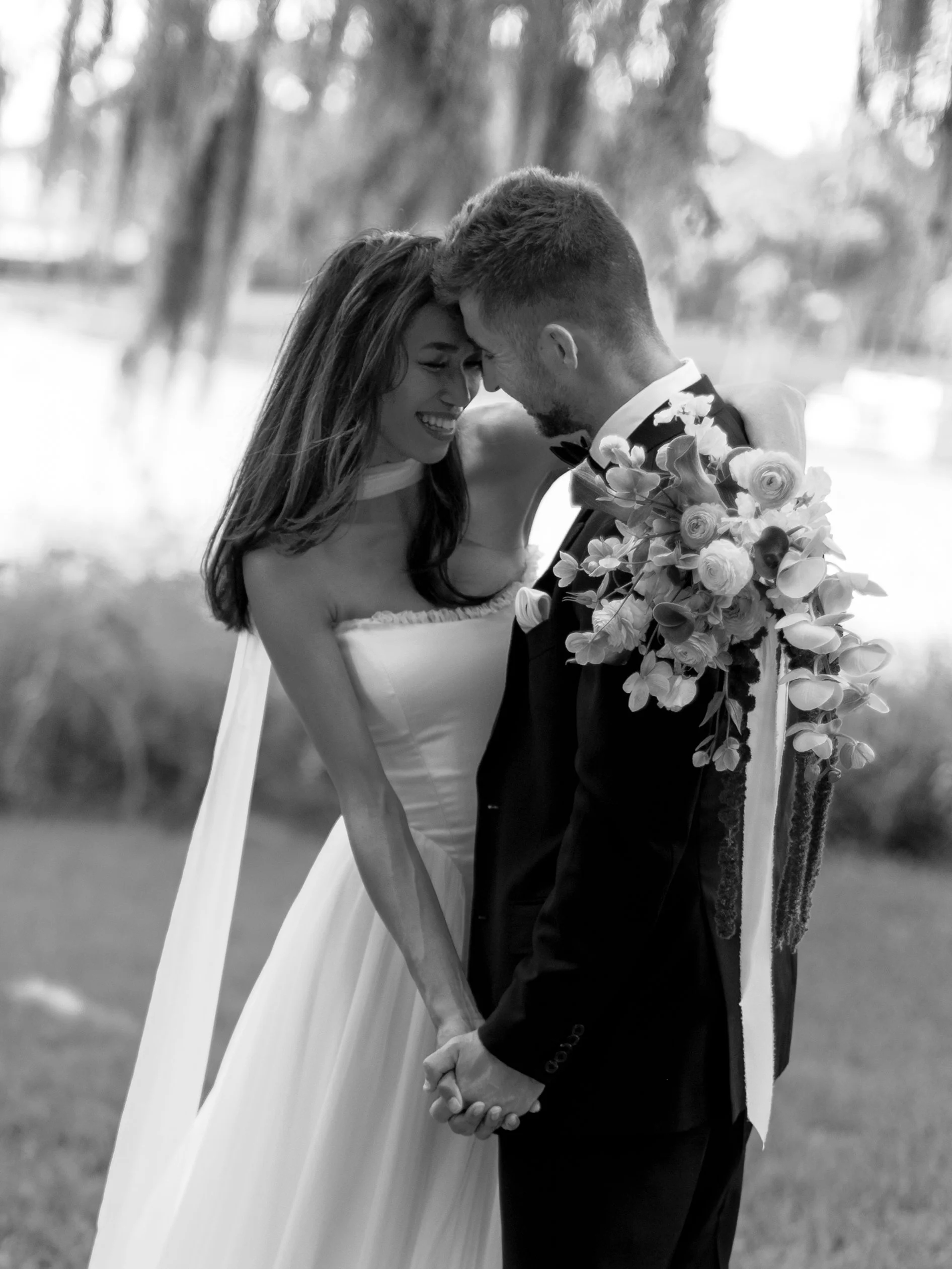 Black and white photo of a bride and groom holding hands, with foreheads touching and smiling, outdoors near a body of water and trees.