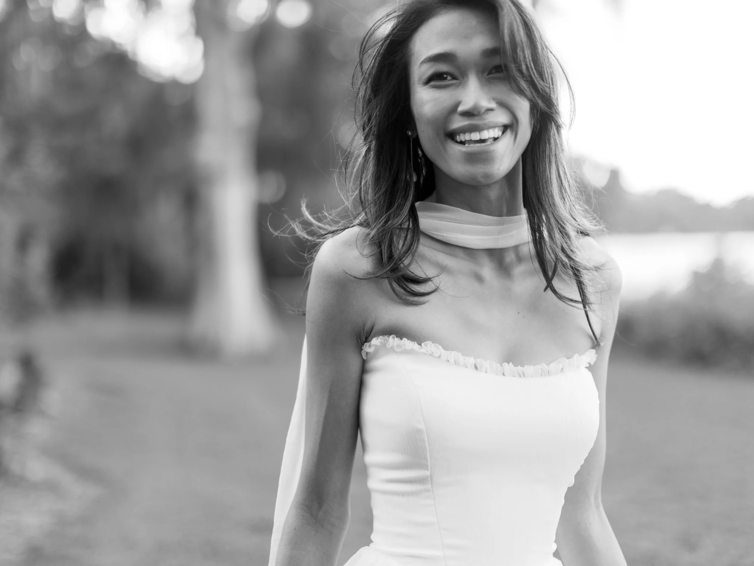 A smiling woman in a strapless dress outdoors in black and white, with trees and a body of water in the background.