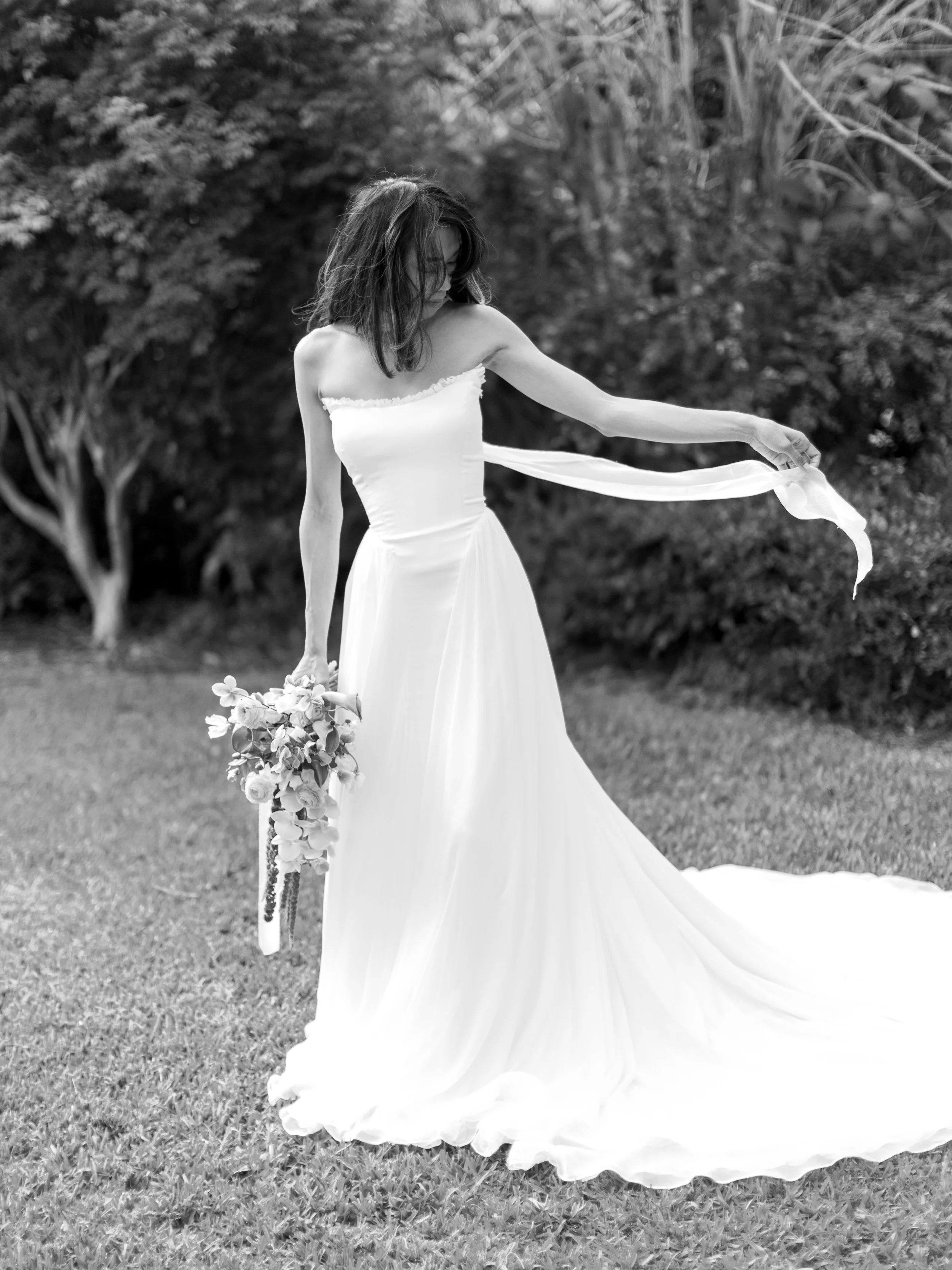 A woman in a white strapless wedding dress holding a bouquet of flowers in a grassy outdoor setting, with her head tilted down and her hair partially covering her face.