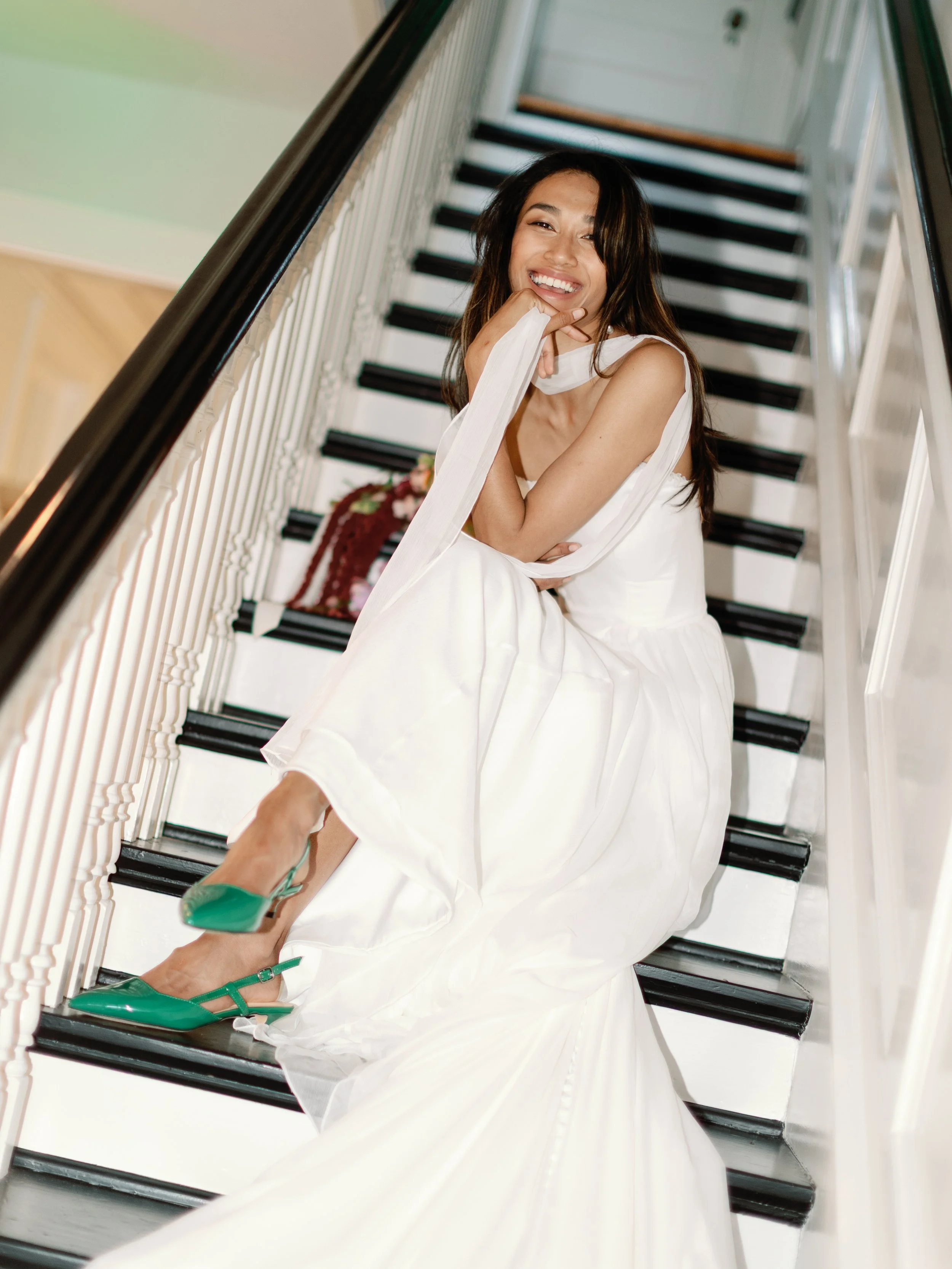 A smiling woman in a white dress and green shoes sitting on a staircase, touching her face with her hand.