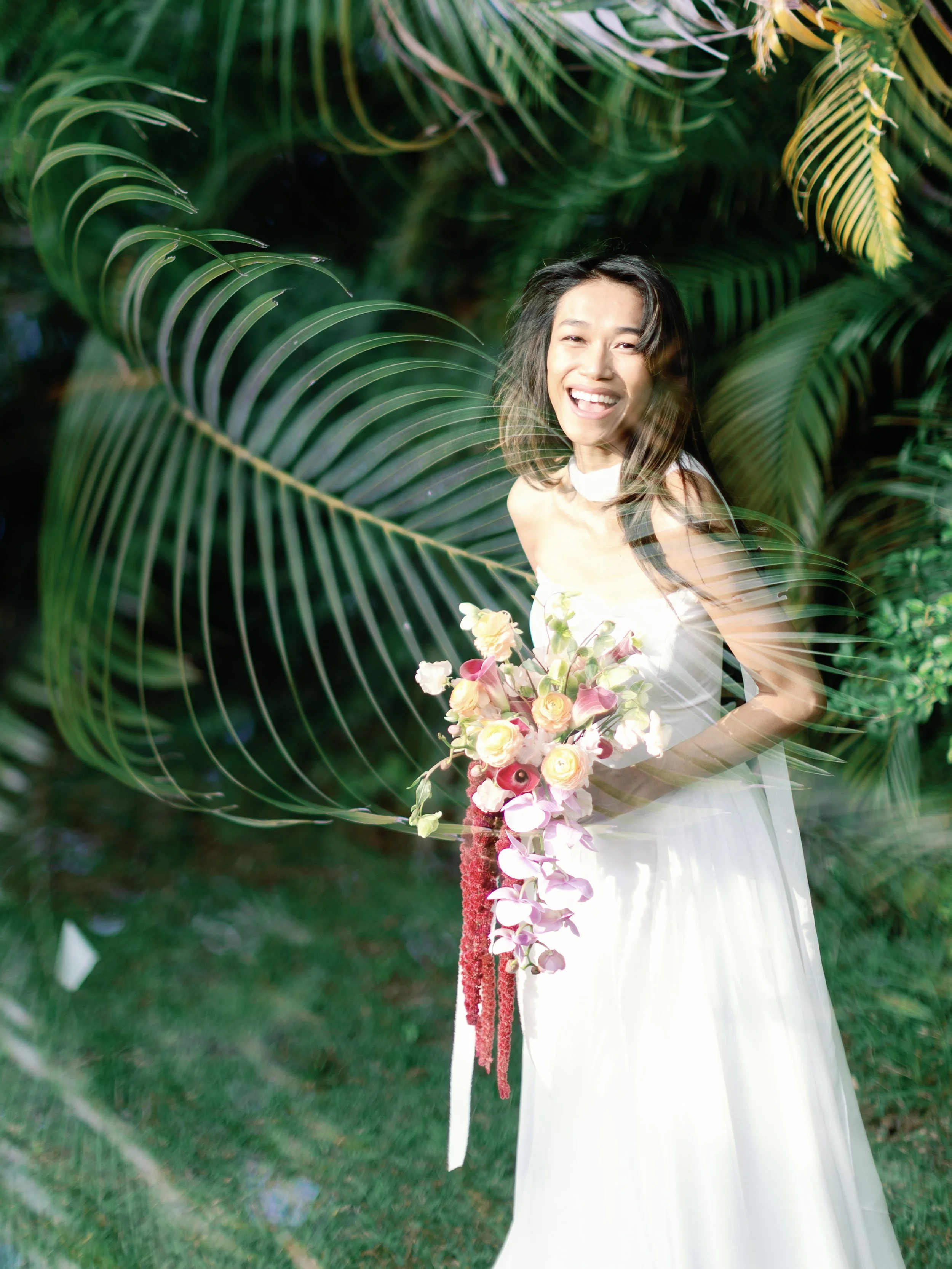 A woman in a white dress holding a bouquet of pink and white flowers standing outdoors with lush green foliage in the background.