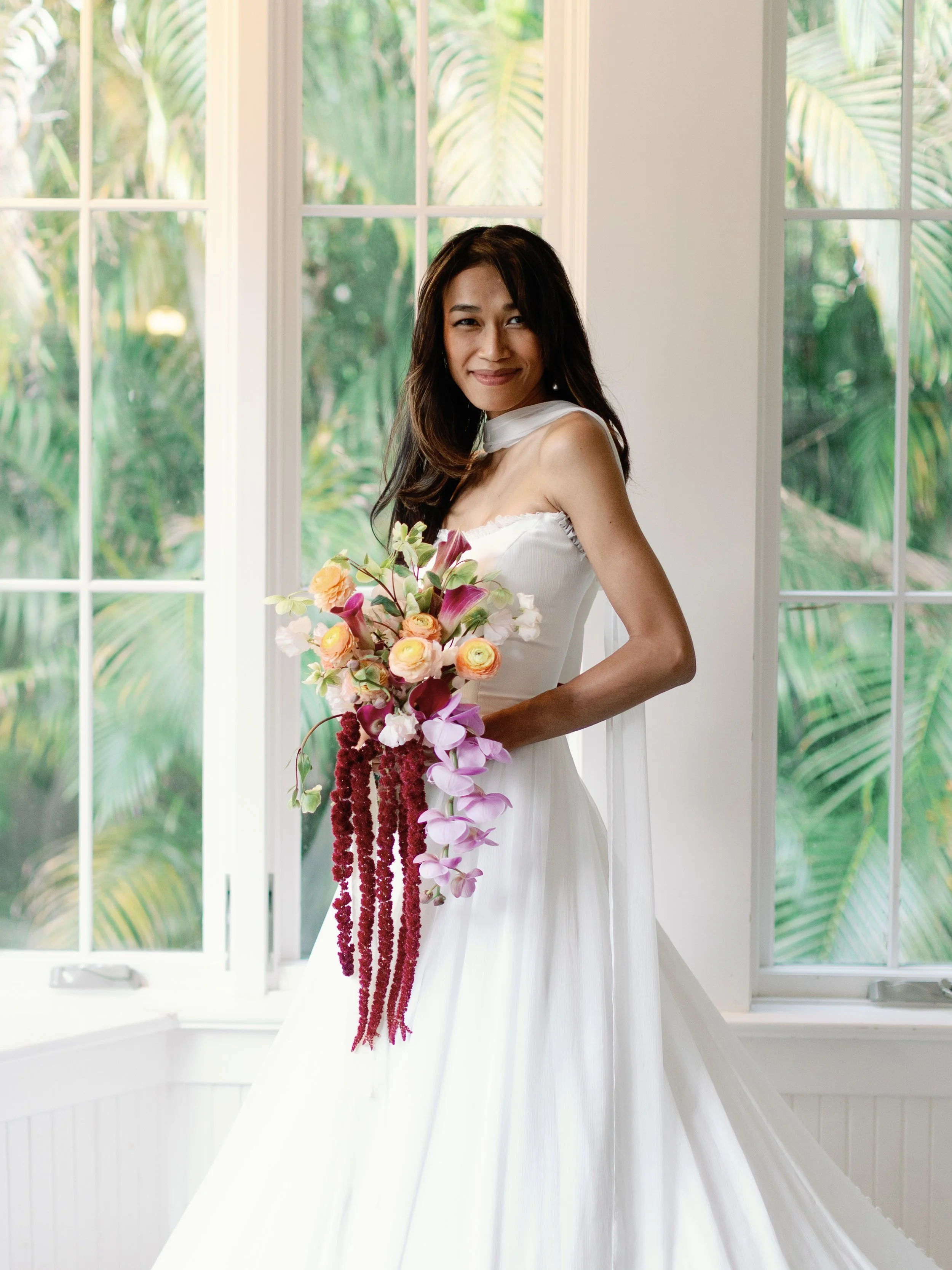 A woman in a white wedding dress holding a cascading bouquet of pink, peach, and purple flowers, standing indoors by large windows with green foliage outside.