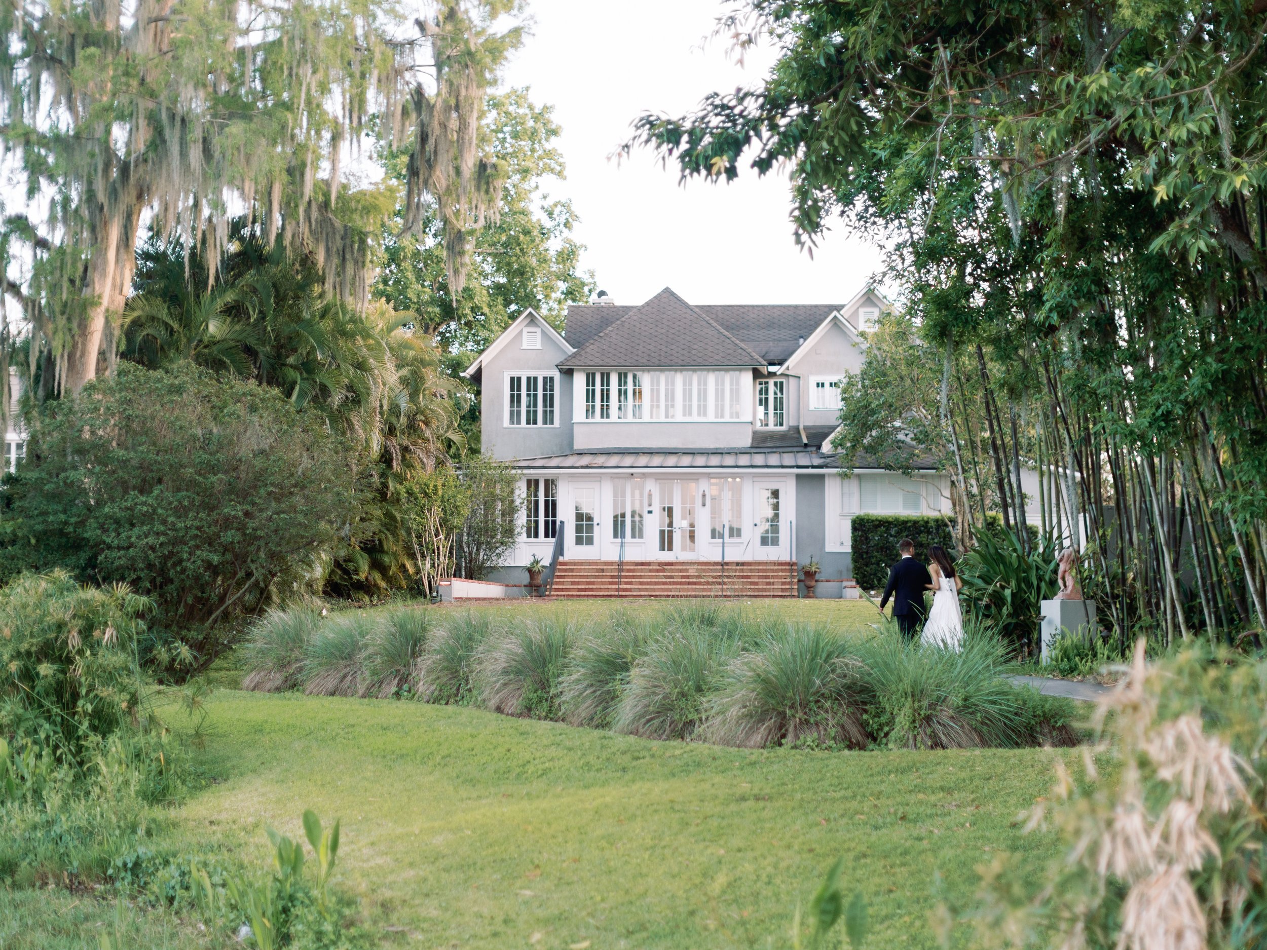 A large gray mansion with white trim and a wide staircase in the backyard, surrounded by lush greenery and trees, with a couple in formal attire walking toward the house.