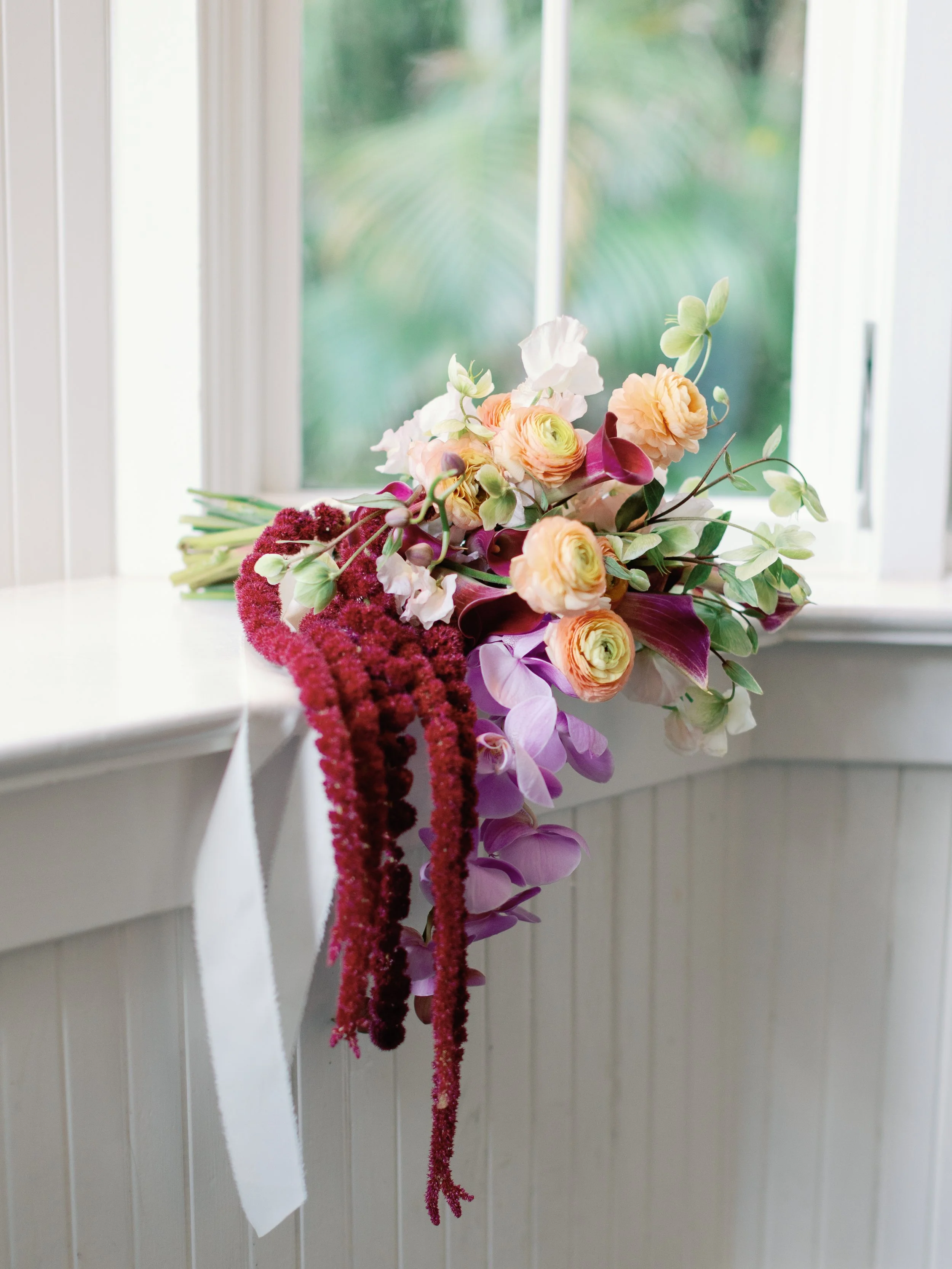 A colorful bouquet of flowers resting on a white windowsill with a window showing green foliage in the background.