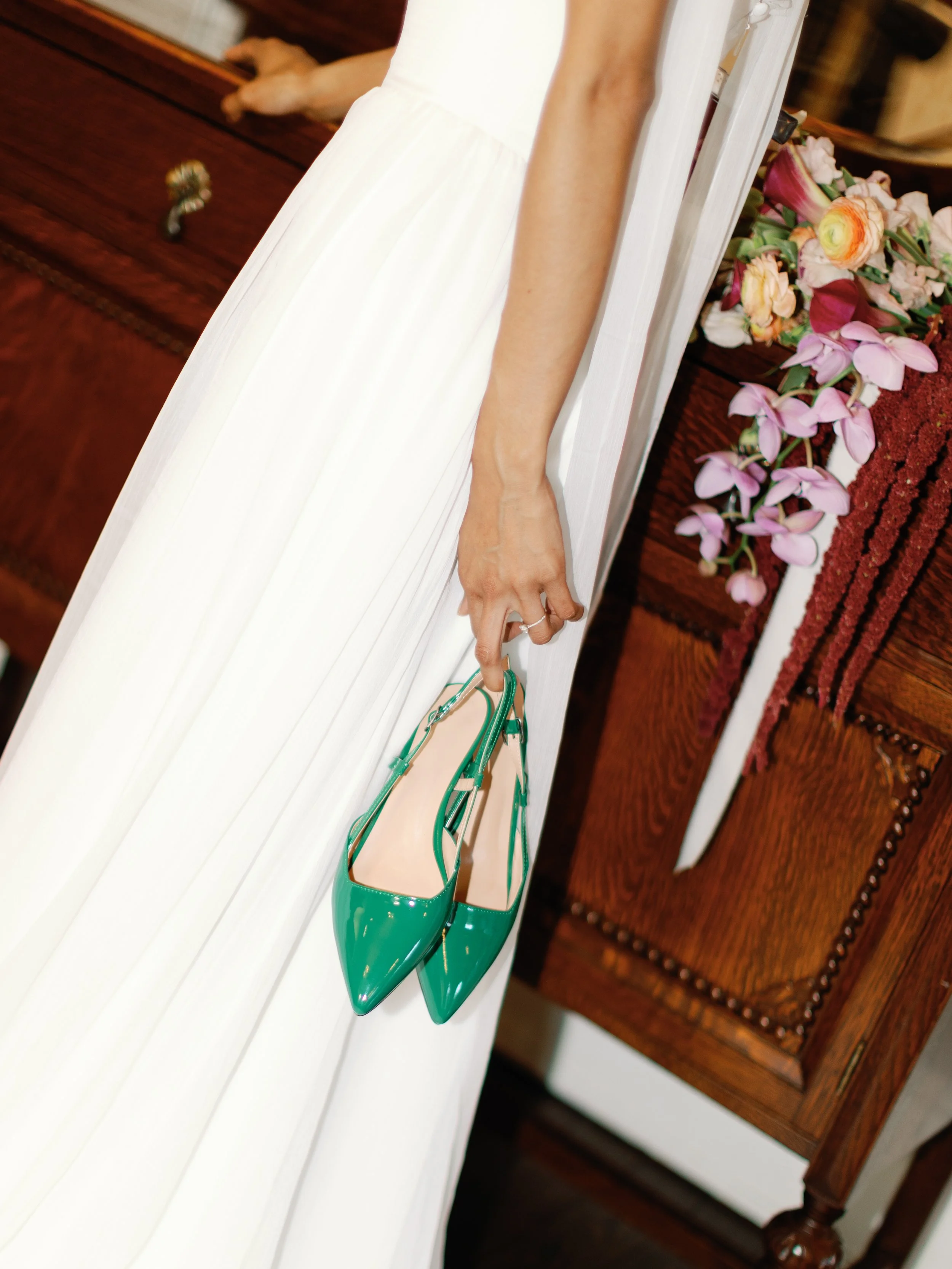 A woman in a white dress holding a pair of green high-heeled shoes with pointed toes, standing next to a wooden table with pink and purple flowers on it.