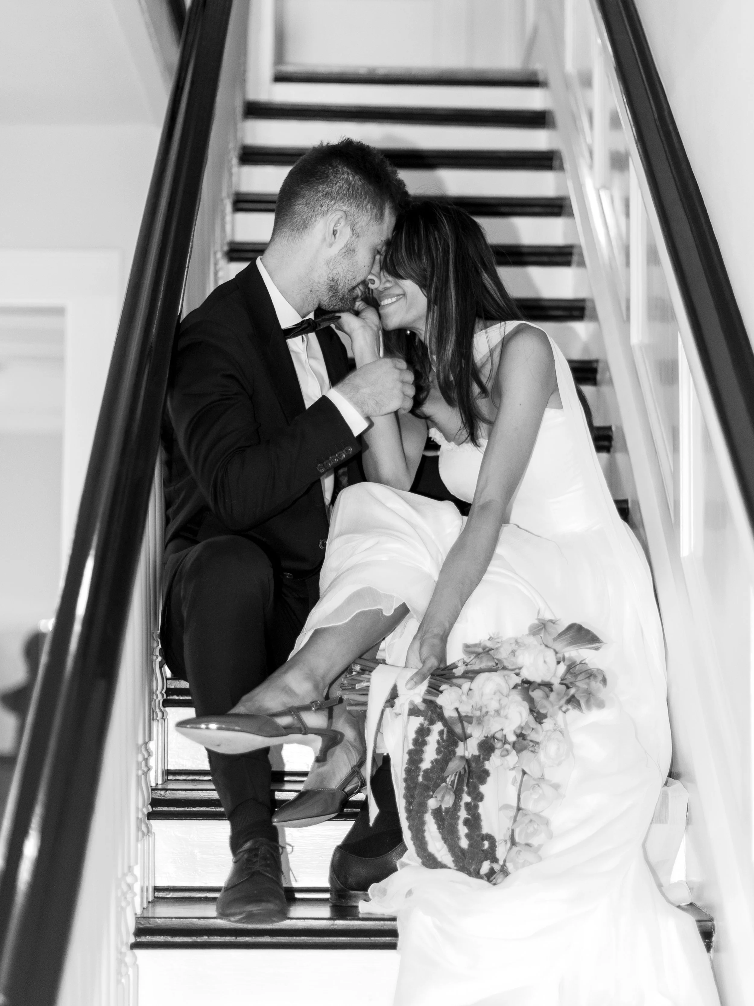 A black and white photo of a couple in wedding attire sitting on stairs. The man is in a tuxedo, and the woman is in a wedding dress holding a bouquet of flowers. They are smiling, touching foreheads, and appear happy.