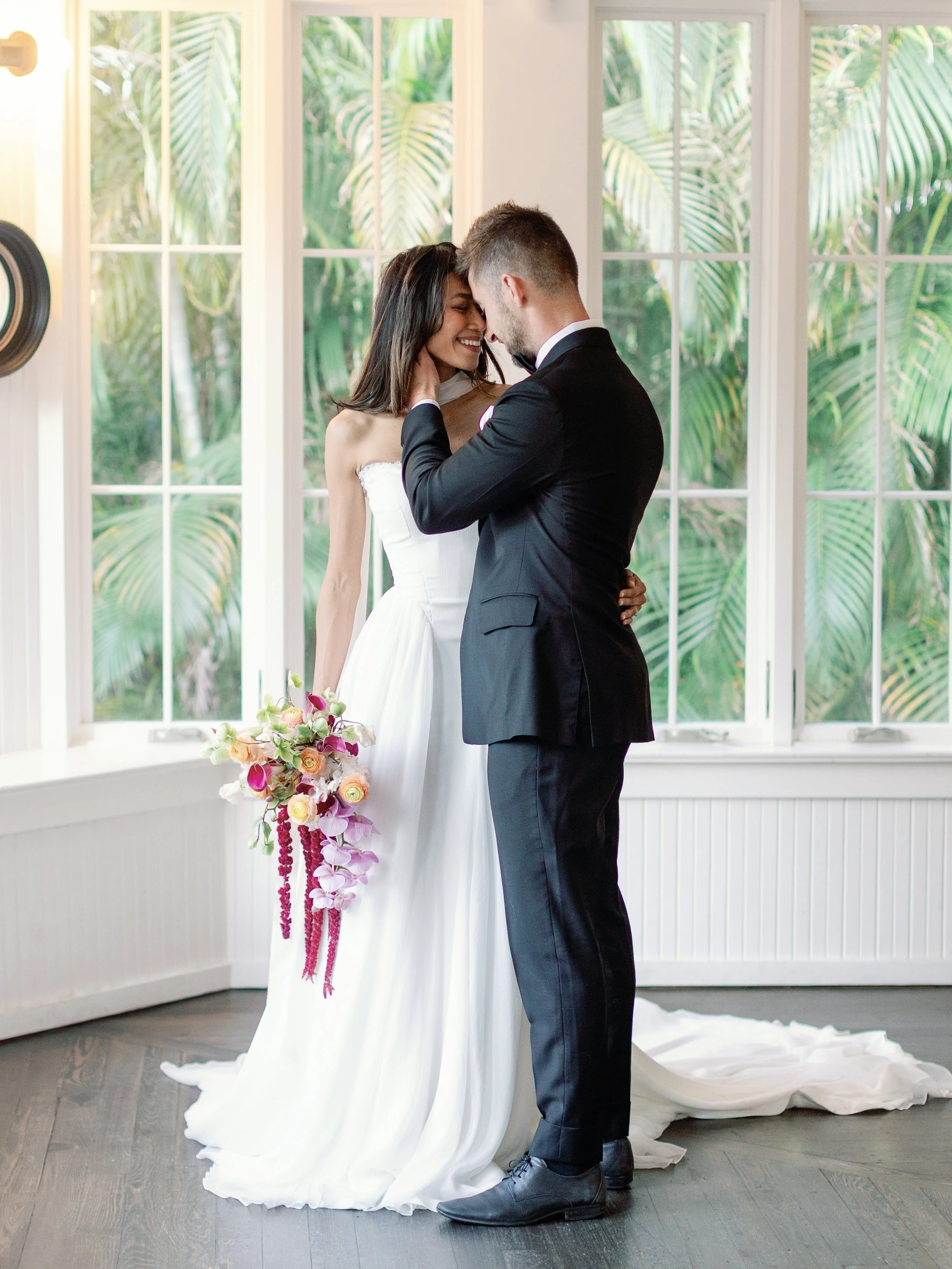 A bride and groom sharing a wedding moment indoors near large windows with green plants outside, with the bride holding a bouquet of pink and purple flowers.