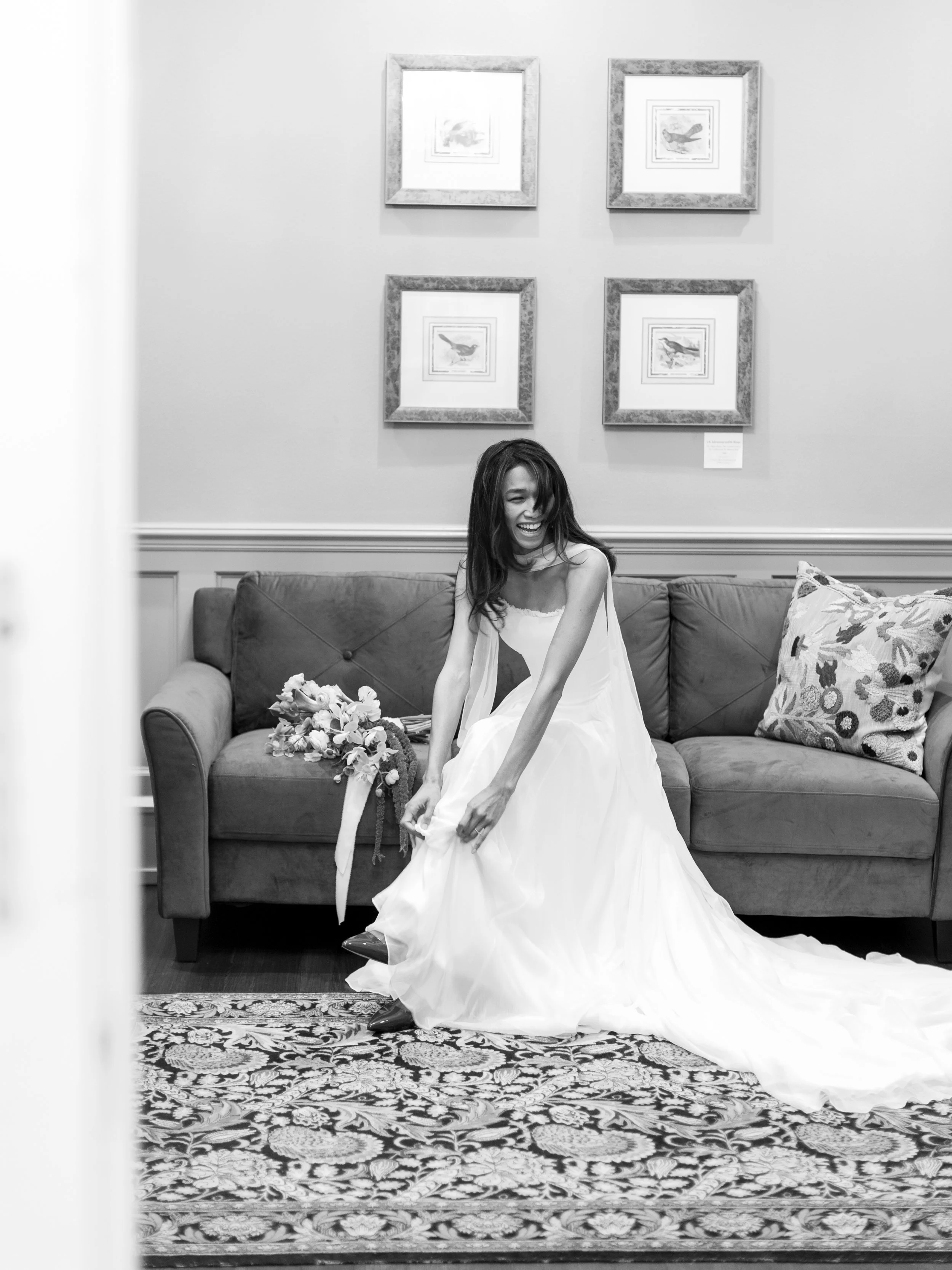 A woman in a wedding dress sitting on a couch, laughing and adjusting her dress. There is a bouquet of flowers on the couch beside her, and framed pictures hang on the wall behind her.