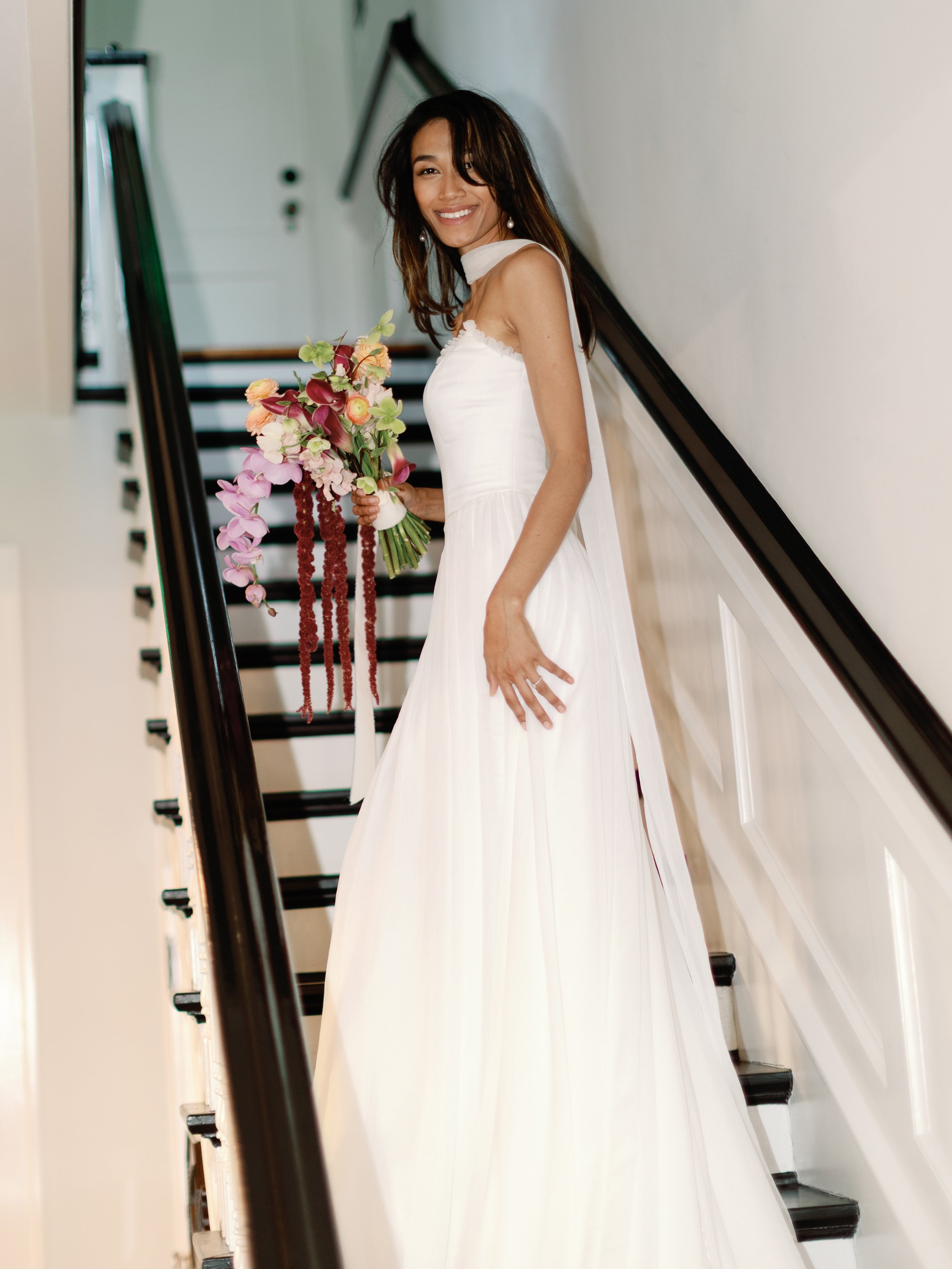 A woman in a white wedding dress holding a bouquet of pink and red flowers, standing on a staircase indoor, smiling.