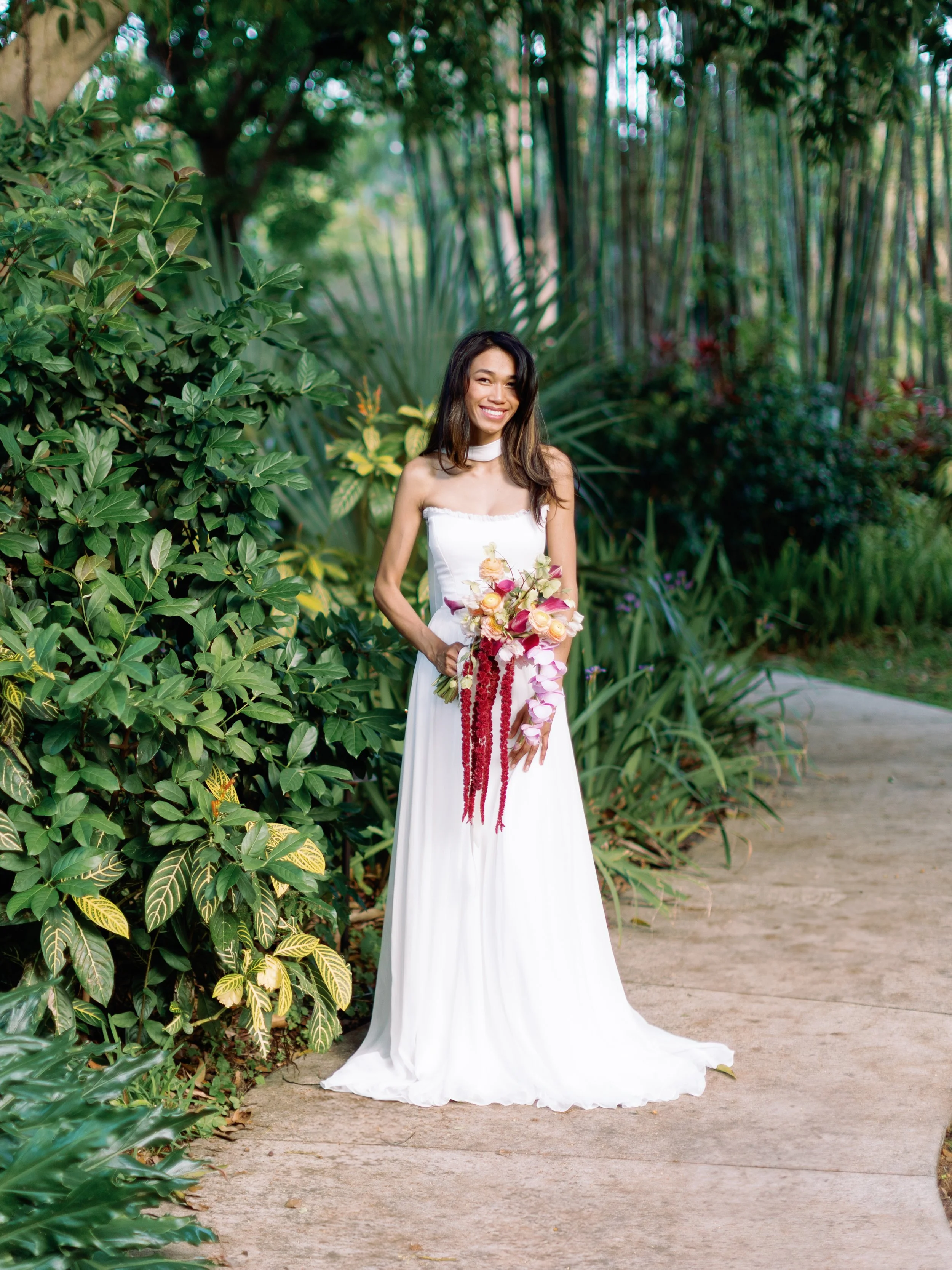 A woman in a white strapless dress holding a colorful bouquet of flowers, standing on a garden path surrounded by lush green foliage.