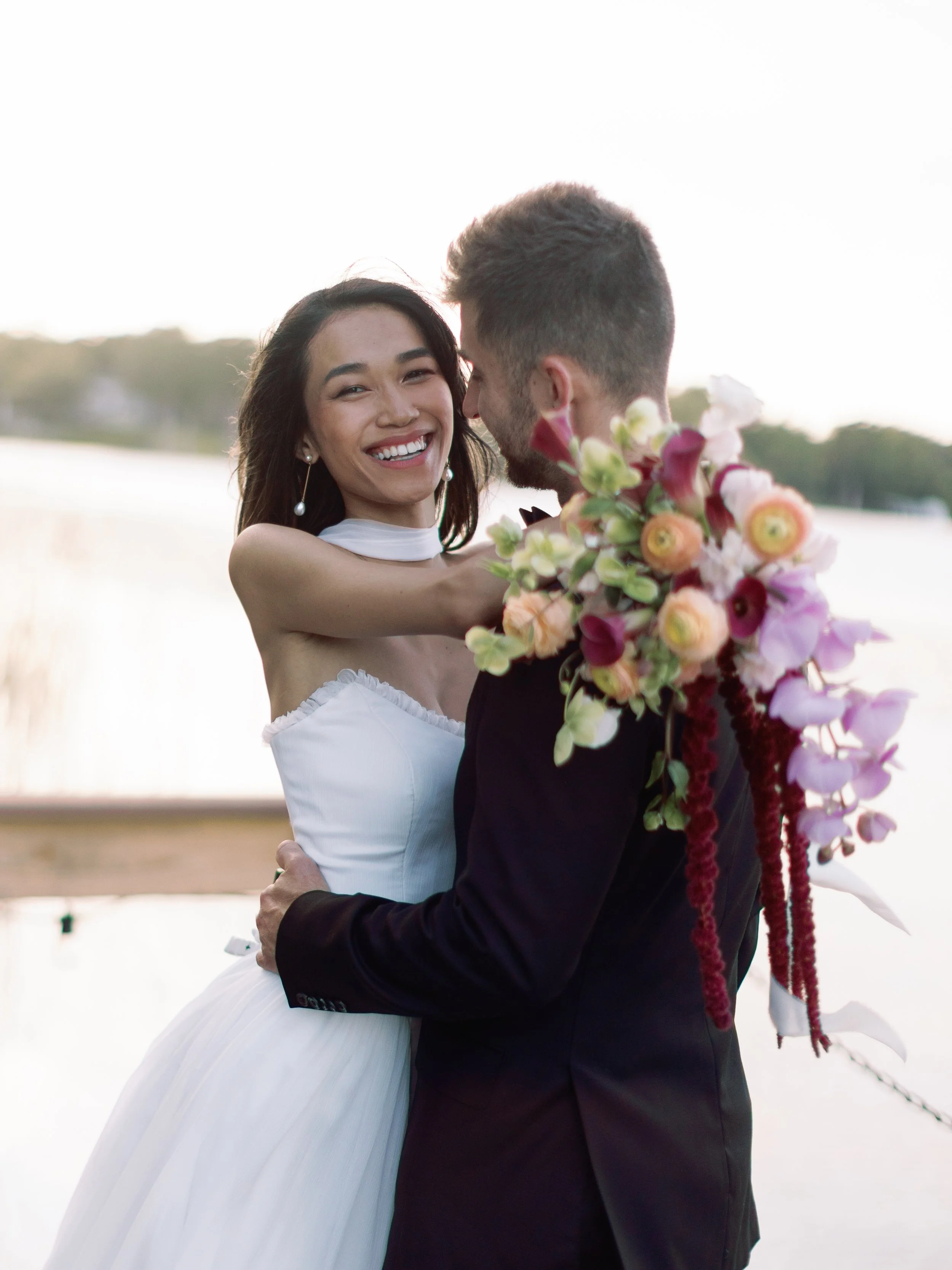 A bride and groom embrace outdoors, the bride wearing a white wedding dress and smiling, holding a large bouquet of colorful flowers, as they look at each other with a natural landscape and water in the background.
