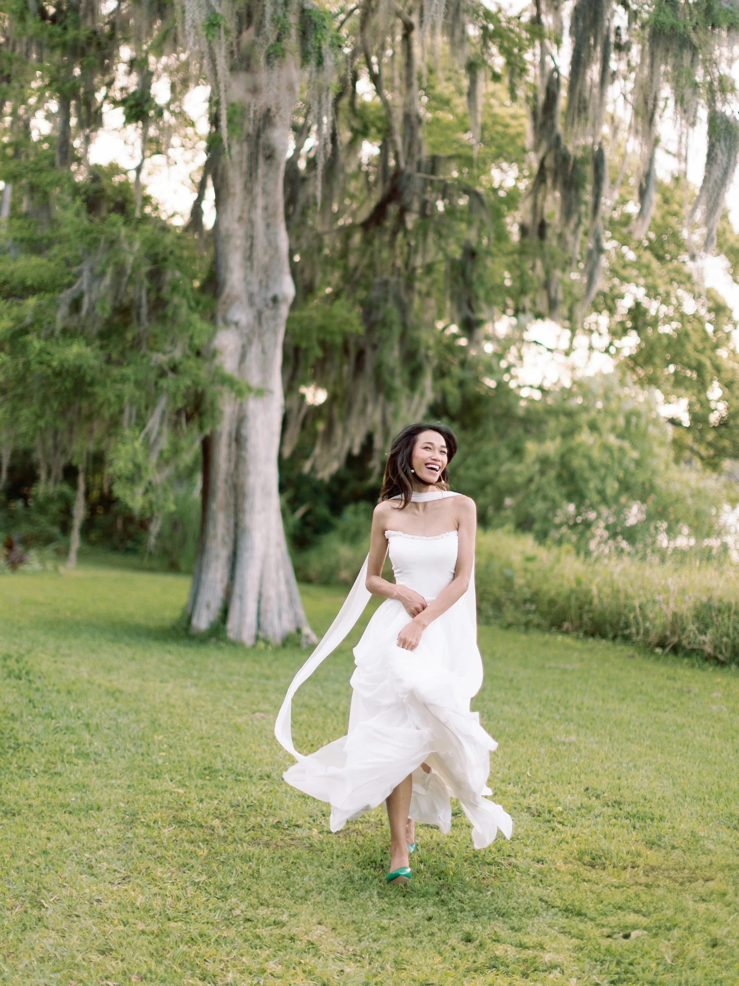 A woman in a white dress running and smiling on a grassy field with trees and moss hanging in the background.