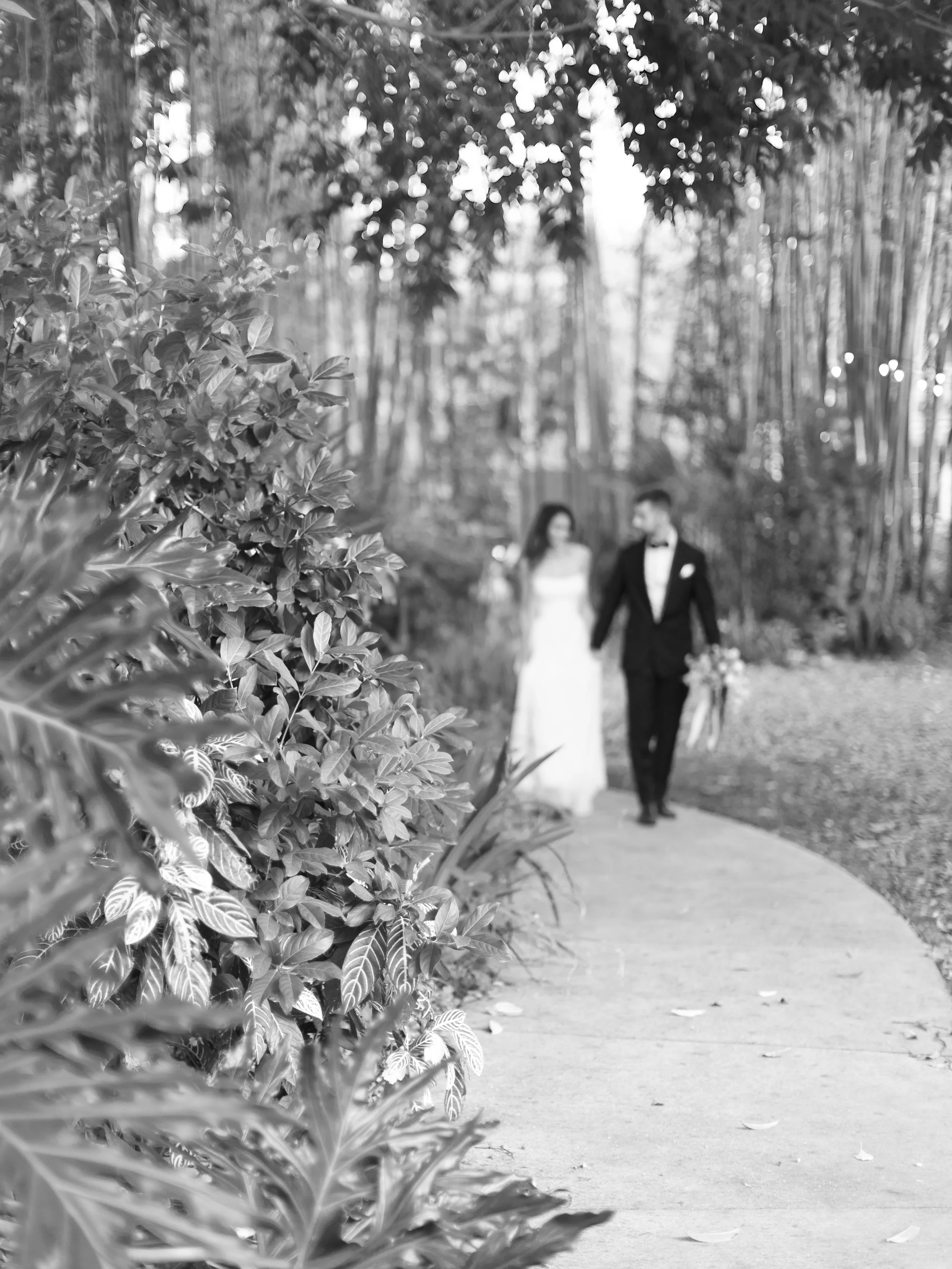 A black and white photo of a bride and groom walking hand in hand along a curved pathway through a garden or park, with trees and foliage in the background.