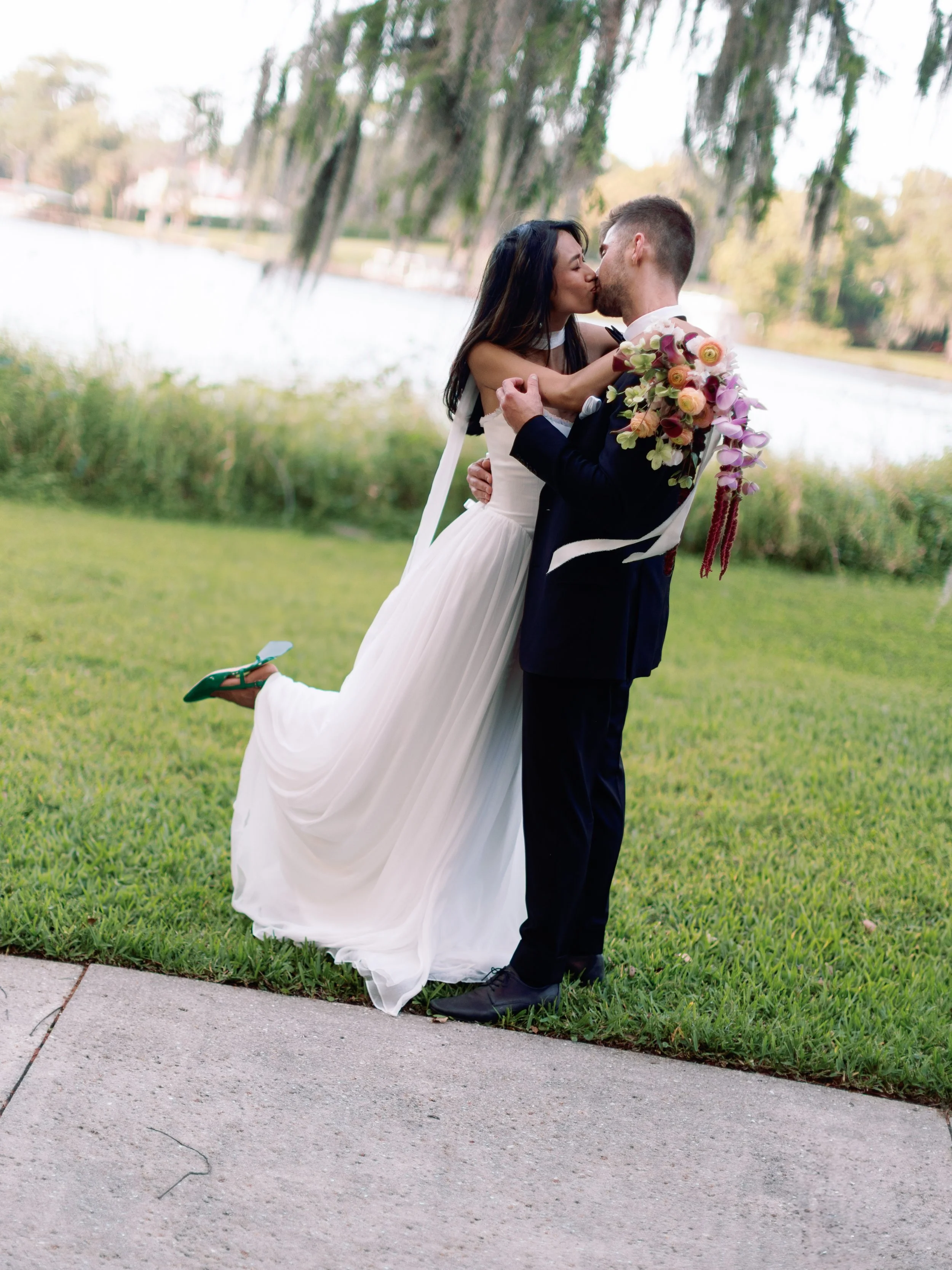 A couple in wedding attire sharing a kiss outdoors near a lake, with the woman in a white gown and the man in a dark suit, holding a colorful bouquet of flowers.