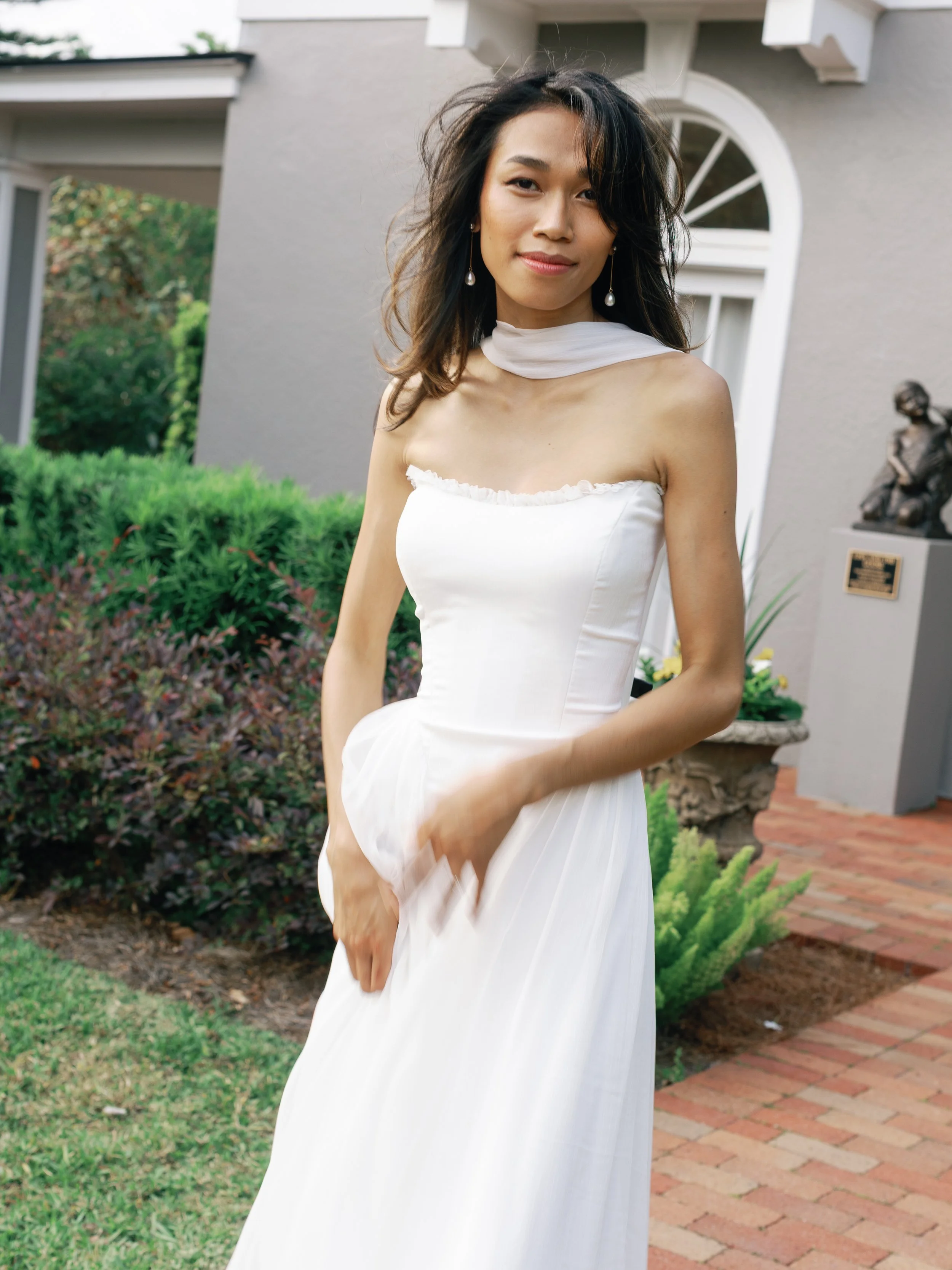 A woman in a white dress standing outdoors near a gray building with lush greenery in the background.