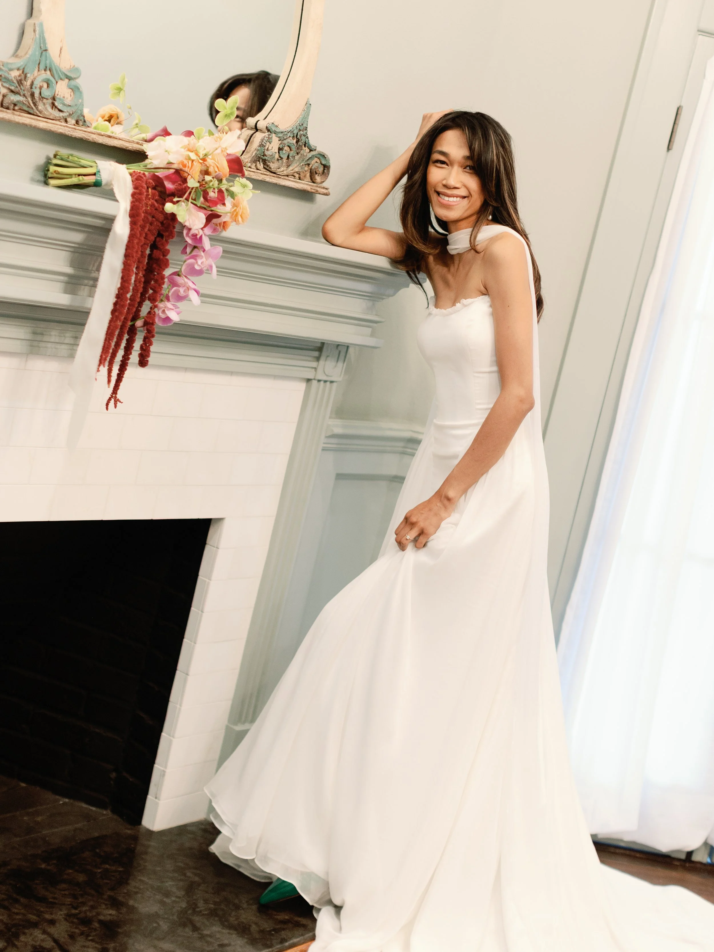 A woman in a white wedding dress smiling and touching her hair, standing next to a decorated fireplace with flowers and a mirror.