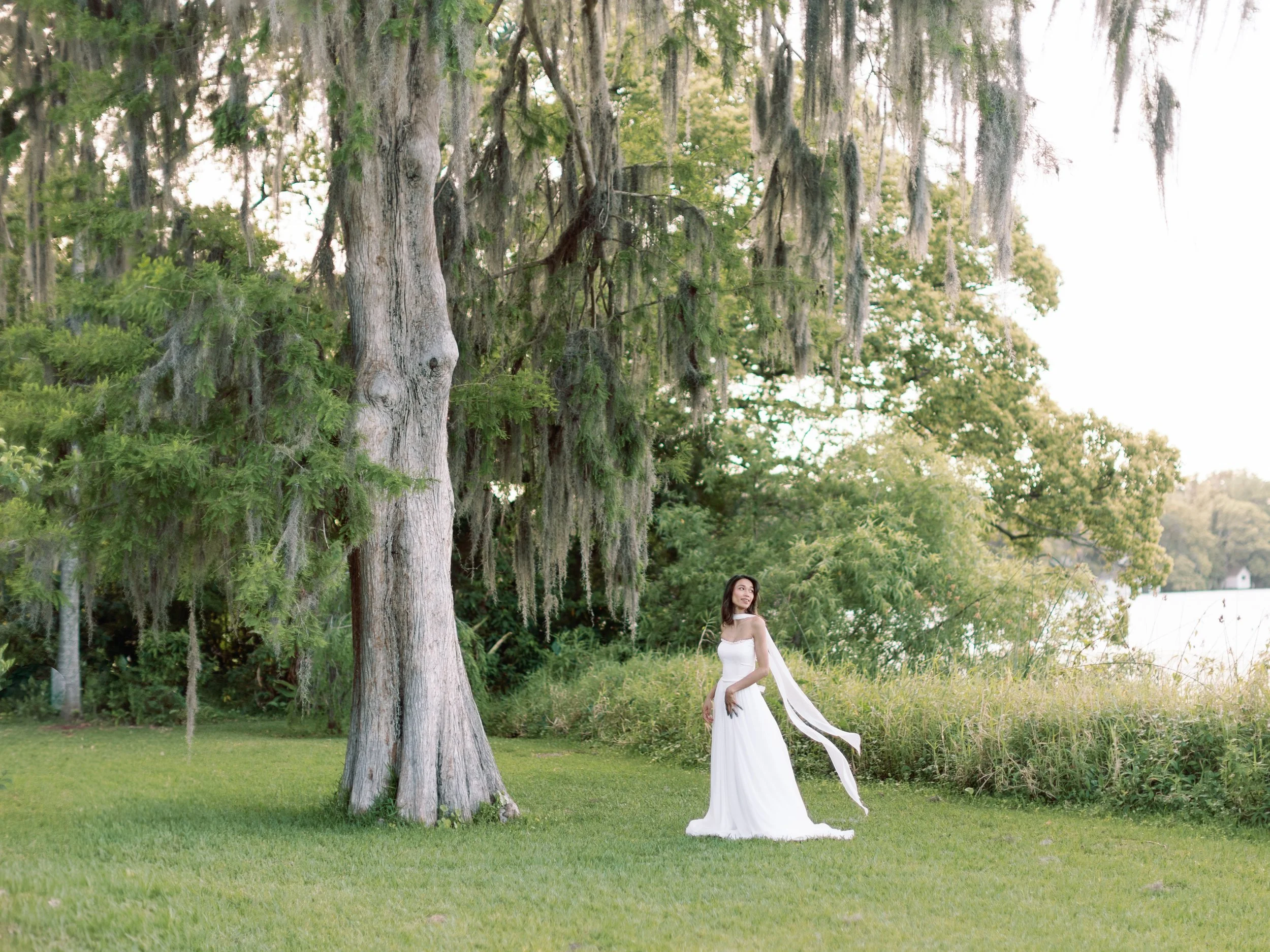 A woman in a white dress standing on a grassy field next to a large tree with hanging moss, in a lush green outdoor setting.