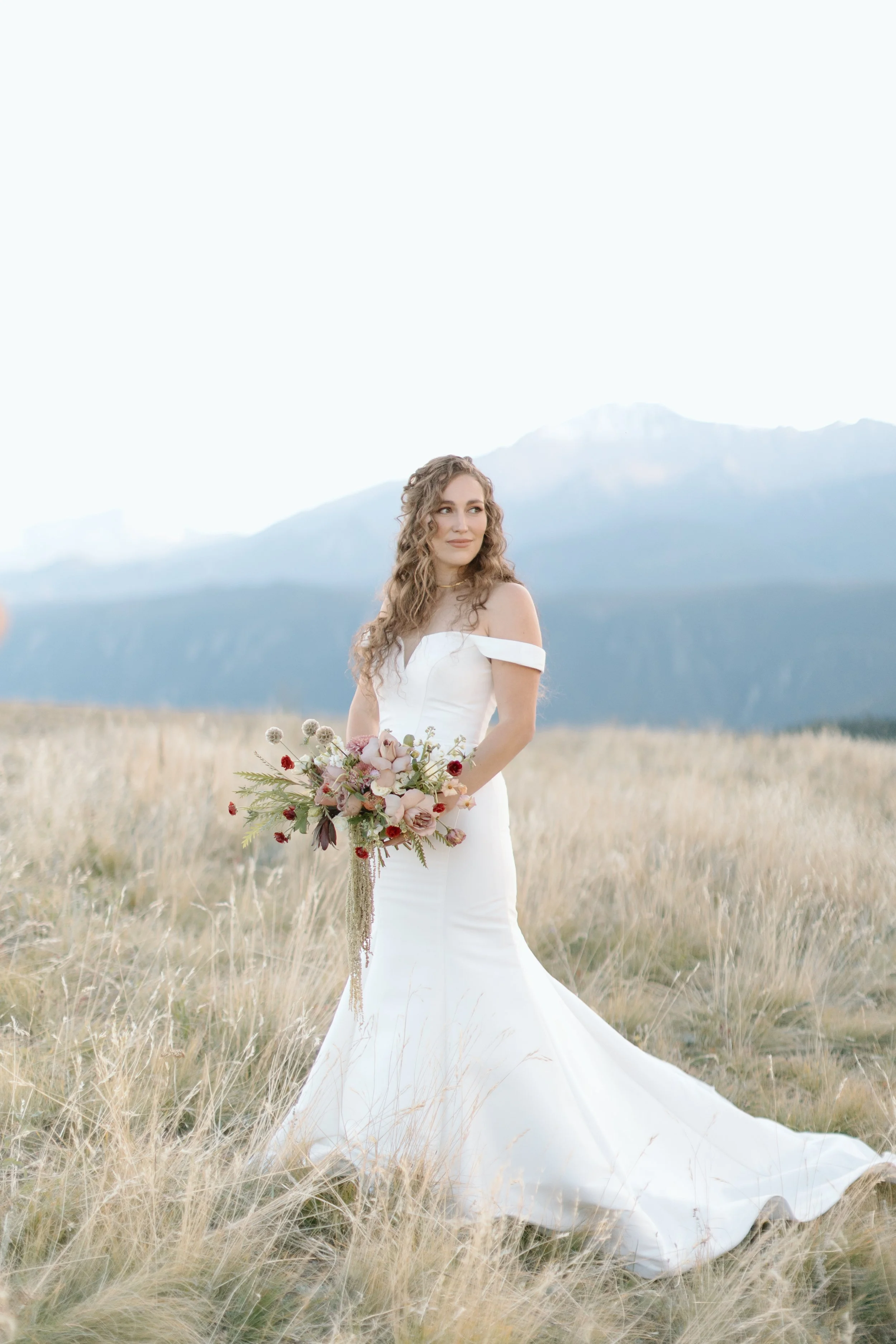 A bride in a white off-shoulder wedding dress holding a bouquet of flowers stands in a grassy field with mountains in the background.
