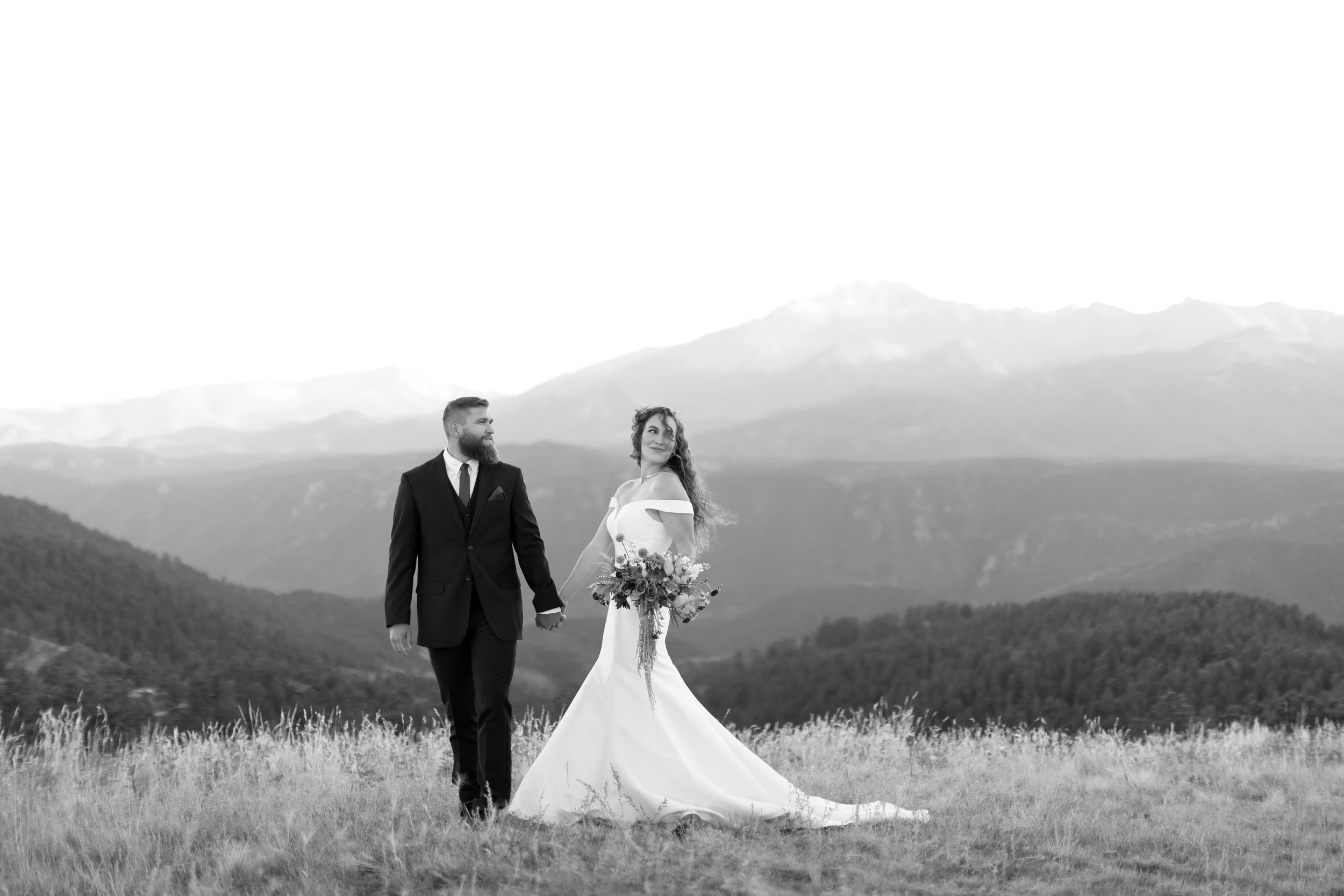 A bride and groom holding hands in a field with mountains in the background, black and white photo.