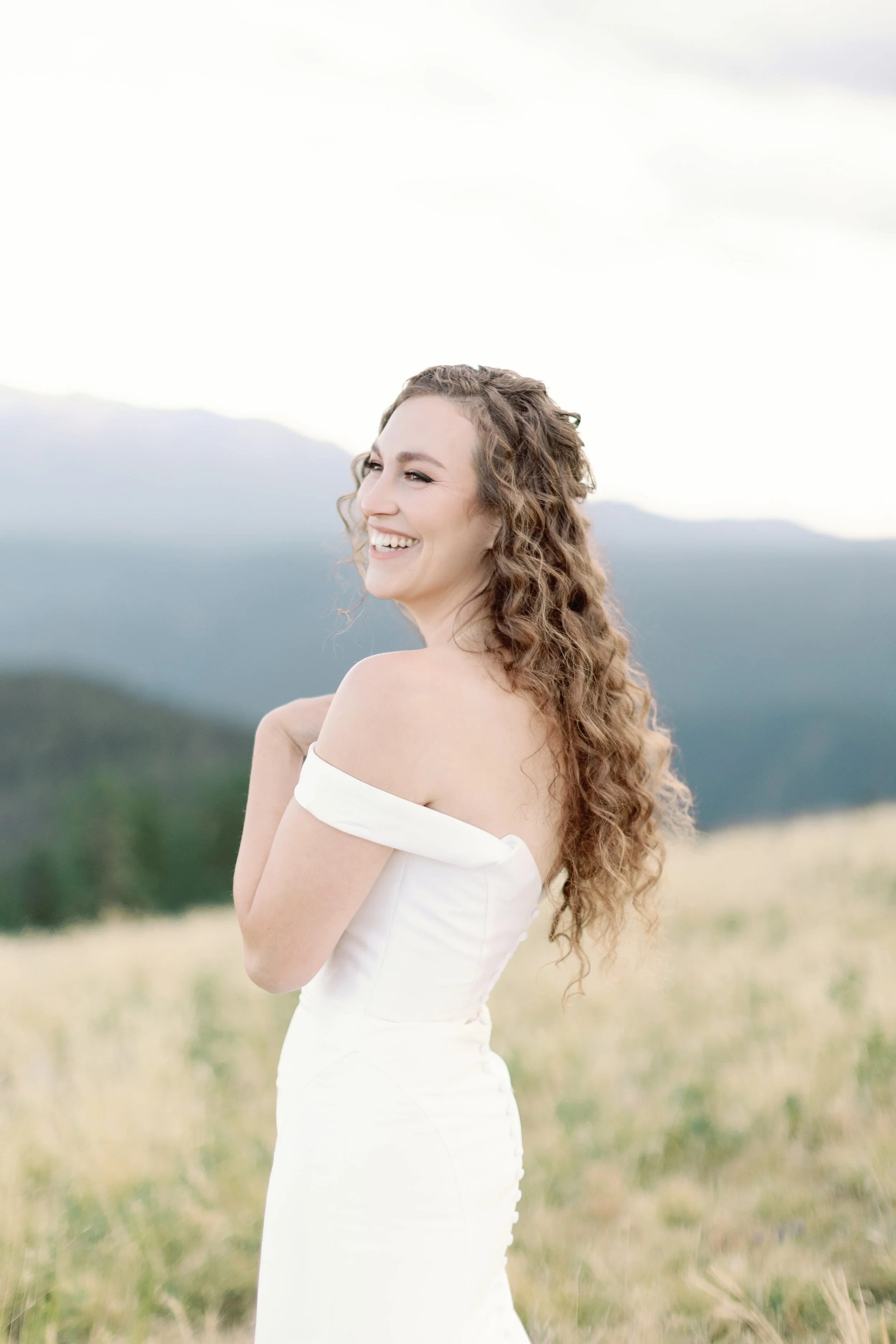A woman in a white off-the-shoulder dress standing in a grassy field with mountains in the background, smiling and looking to her left.