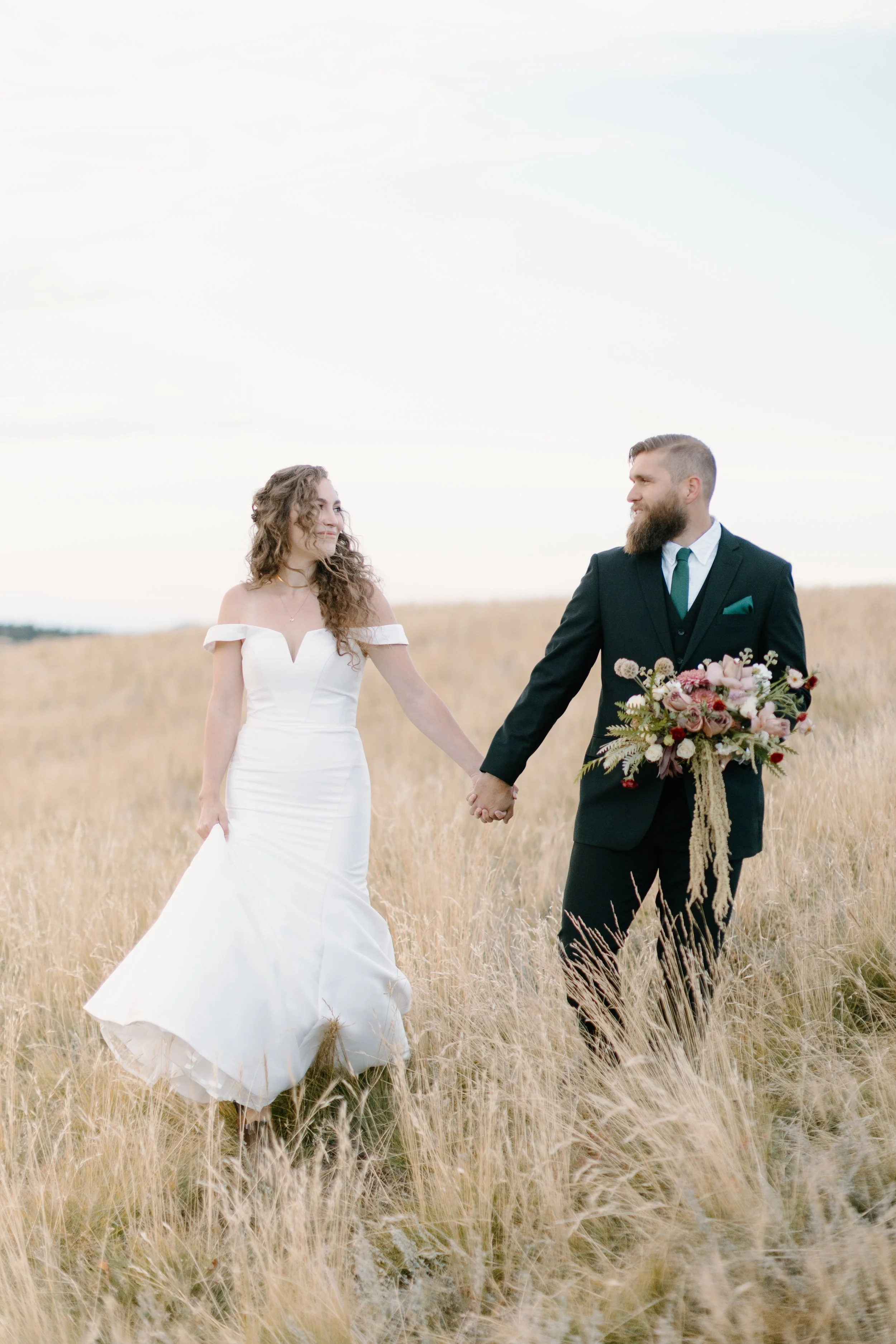 A bride and groom holding hands in a field of tall grass, with the bride in a white off-shoulder wedding dress and the groom in a black suit holding a bouquet of flowers.