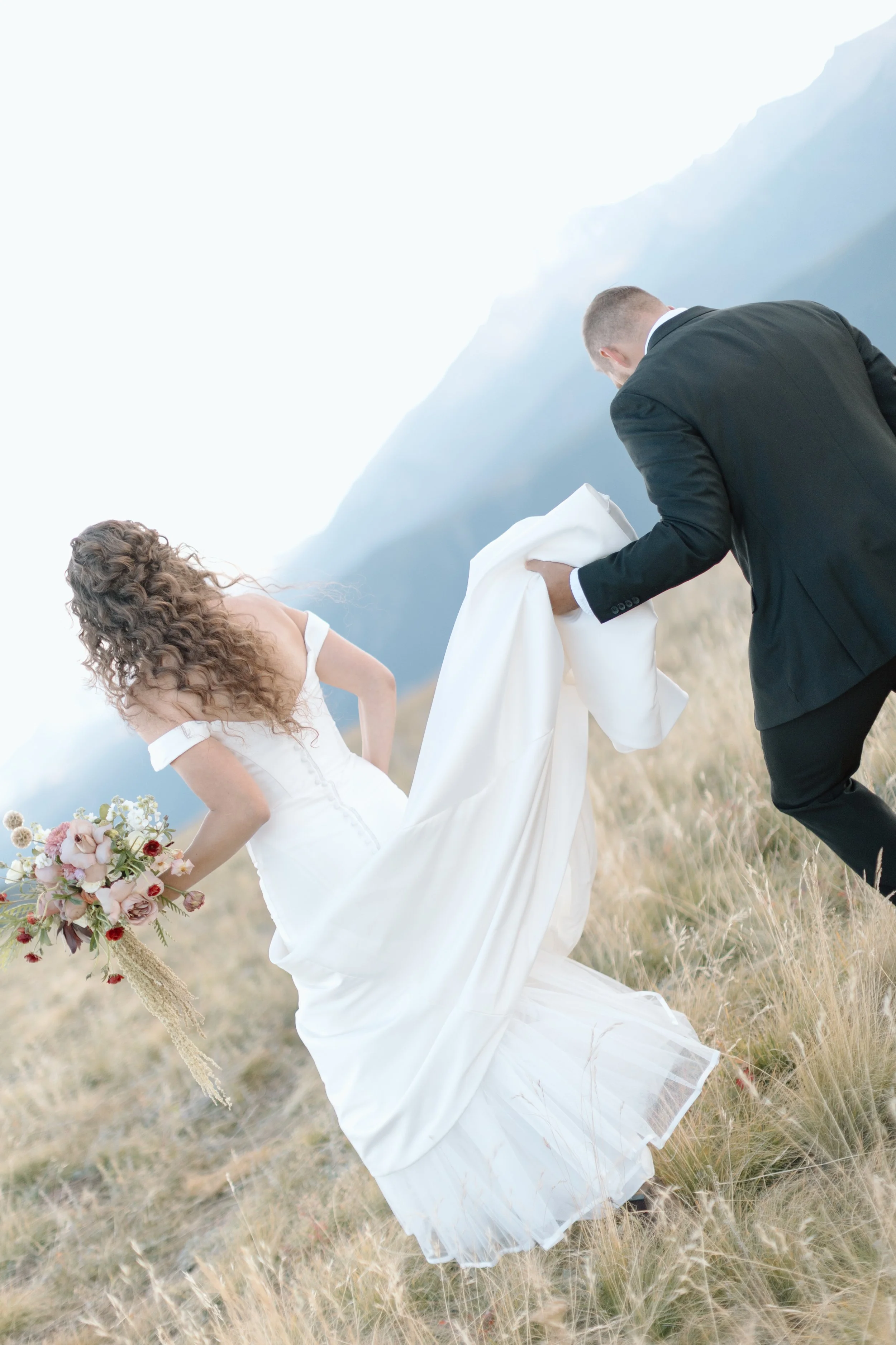 Bride in a white wedding dress holding a bouquet of flowers, running on a grassy field while groom in a black suit helps lift her dress.