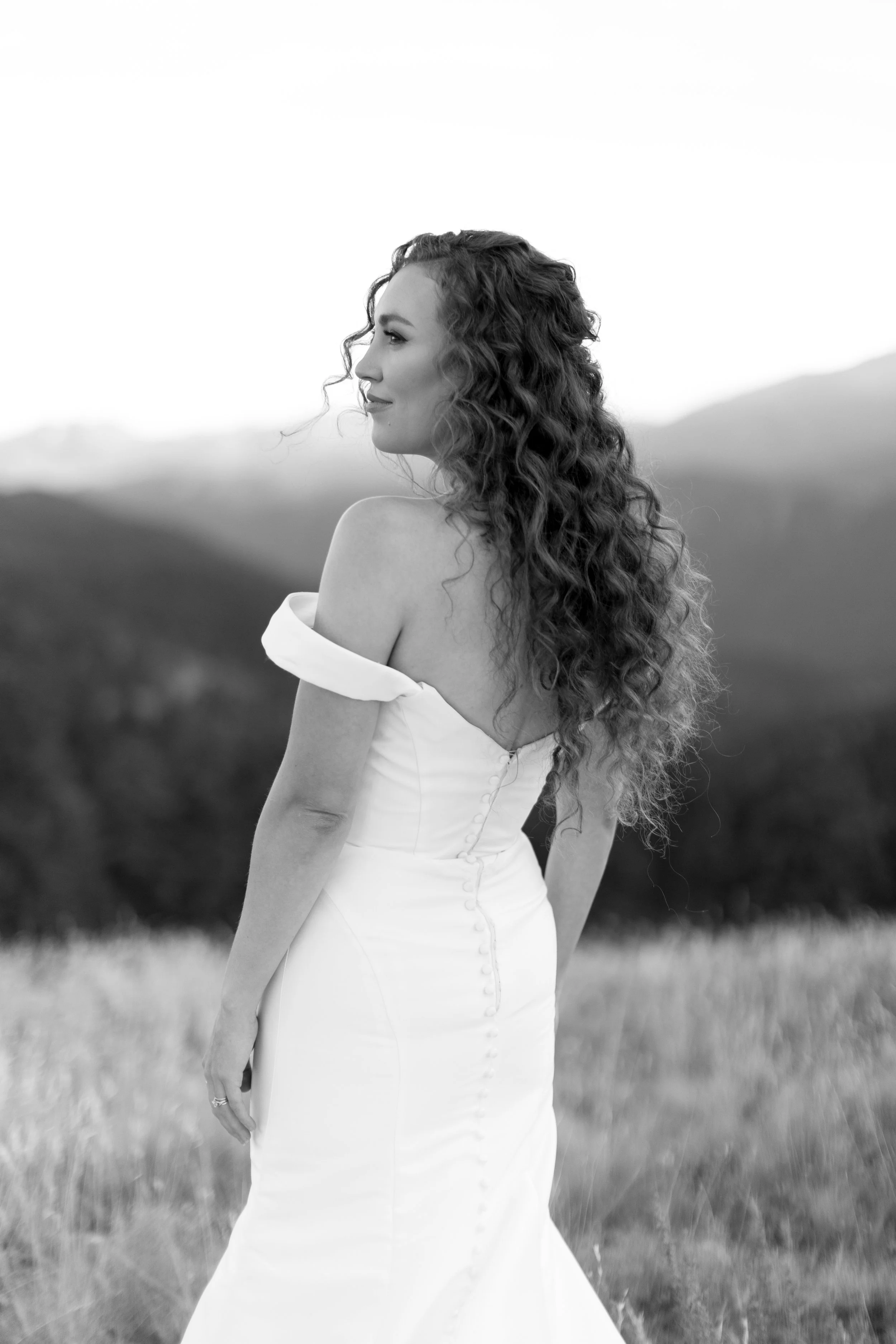 A black and white photo of a bride in a wedding dress standing in a field, looking over her shoulder at the mountains around her