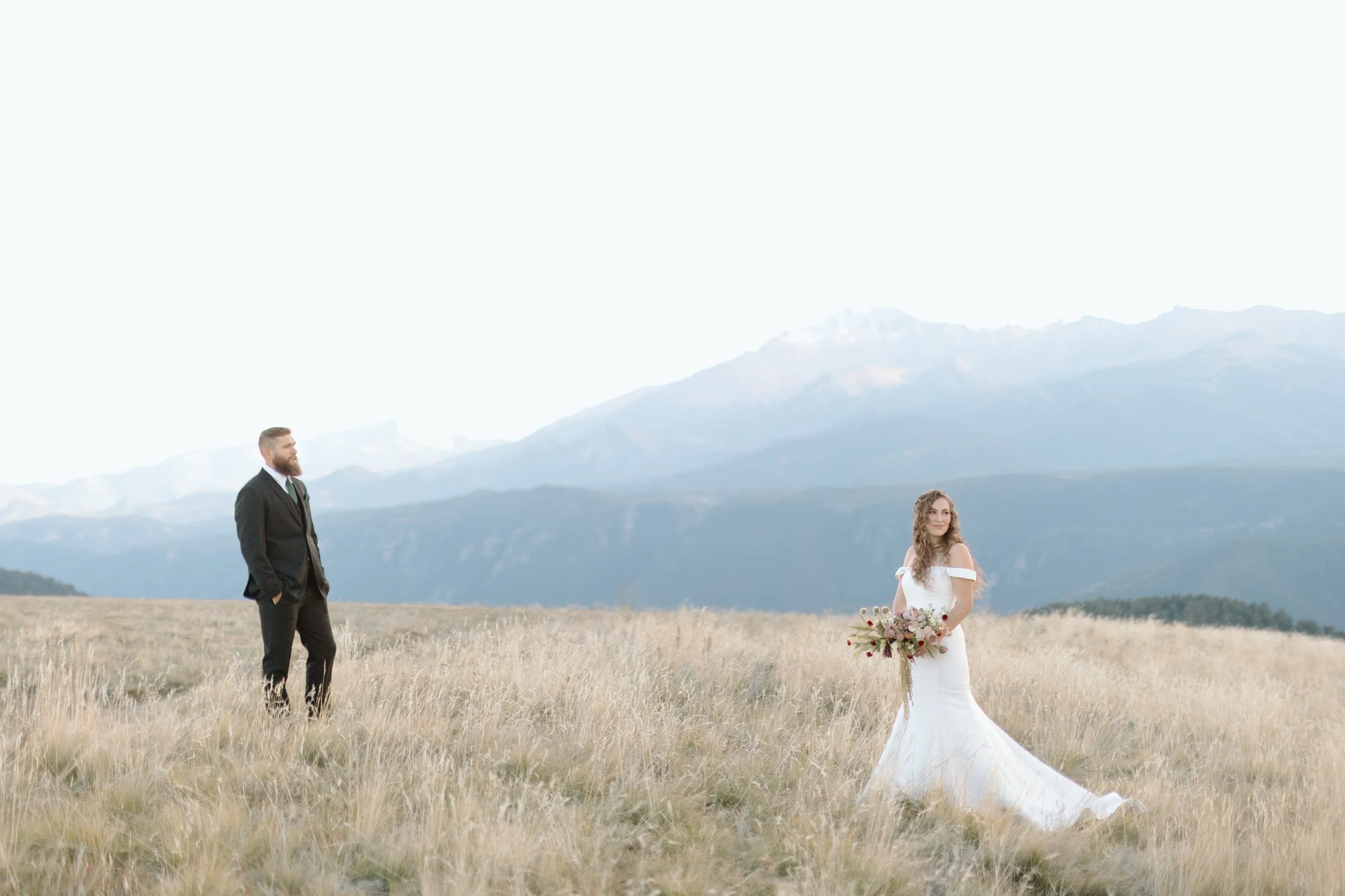 Bride in white wedding dress holding bouquet and groom in black suit standing apart on a grassy field with mountains in the background.