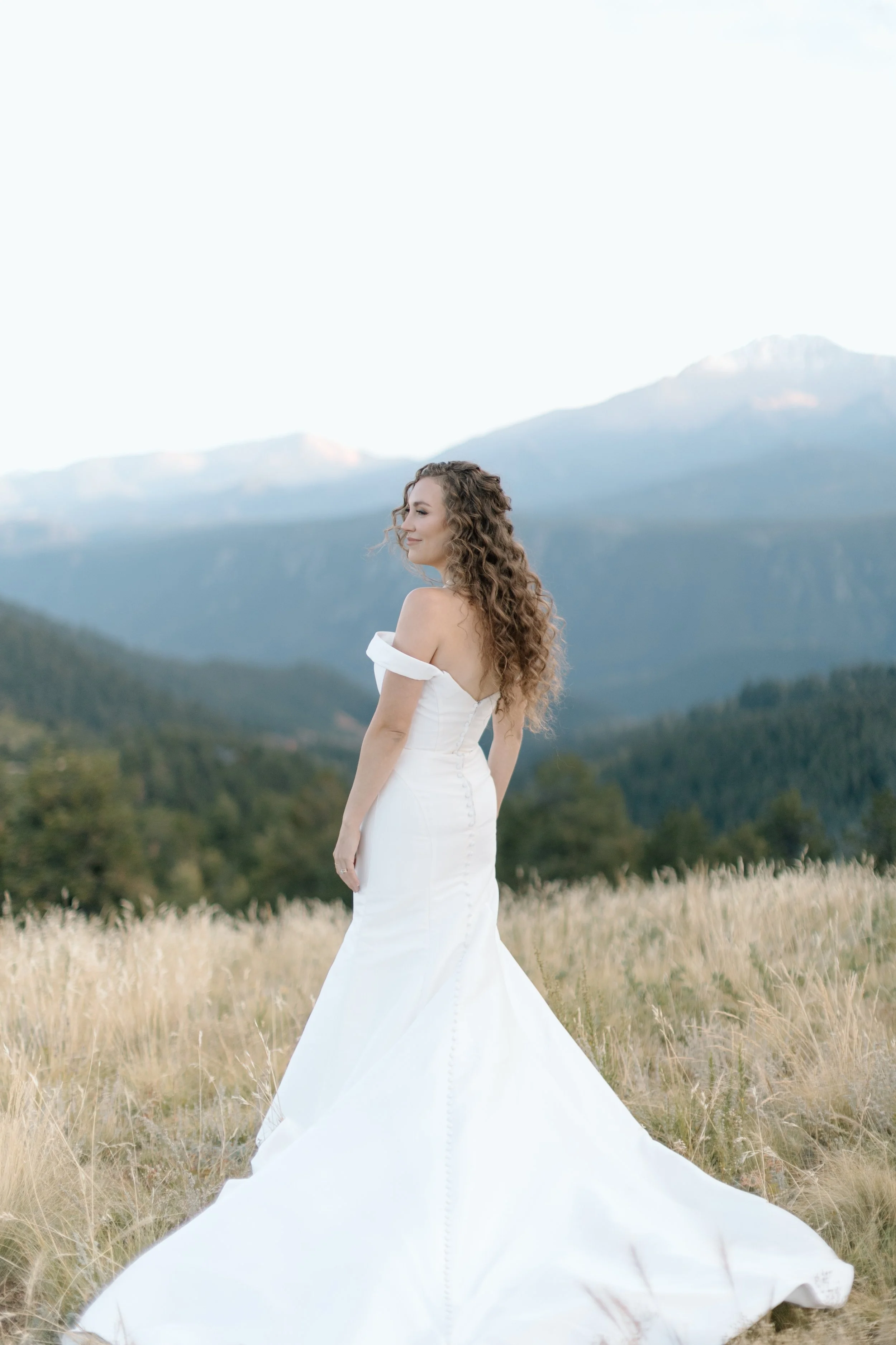 A woman in a white wedding dress standing in a grassy field with mountains and trees in the background.