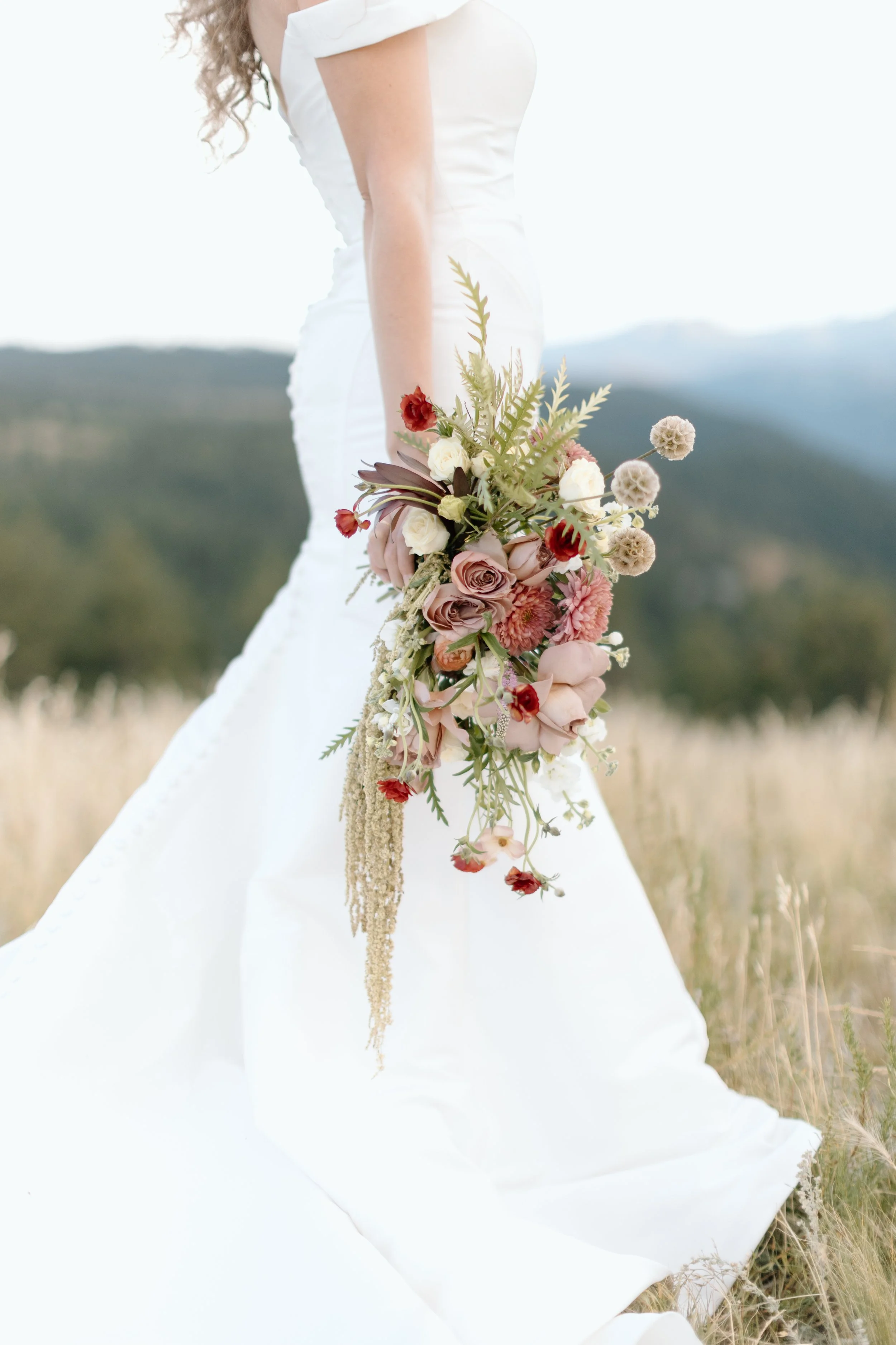 Bride in white wedding dress holding a cascading bouquet of pink, white, and red flowers outdoors in a field.