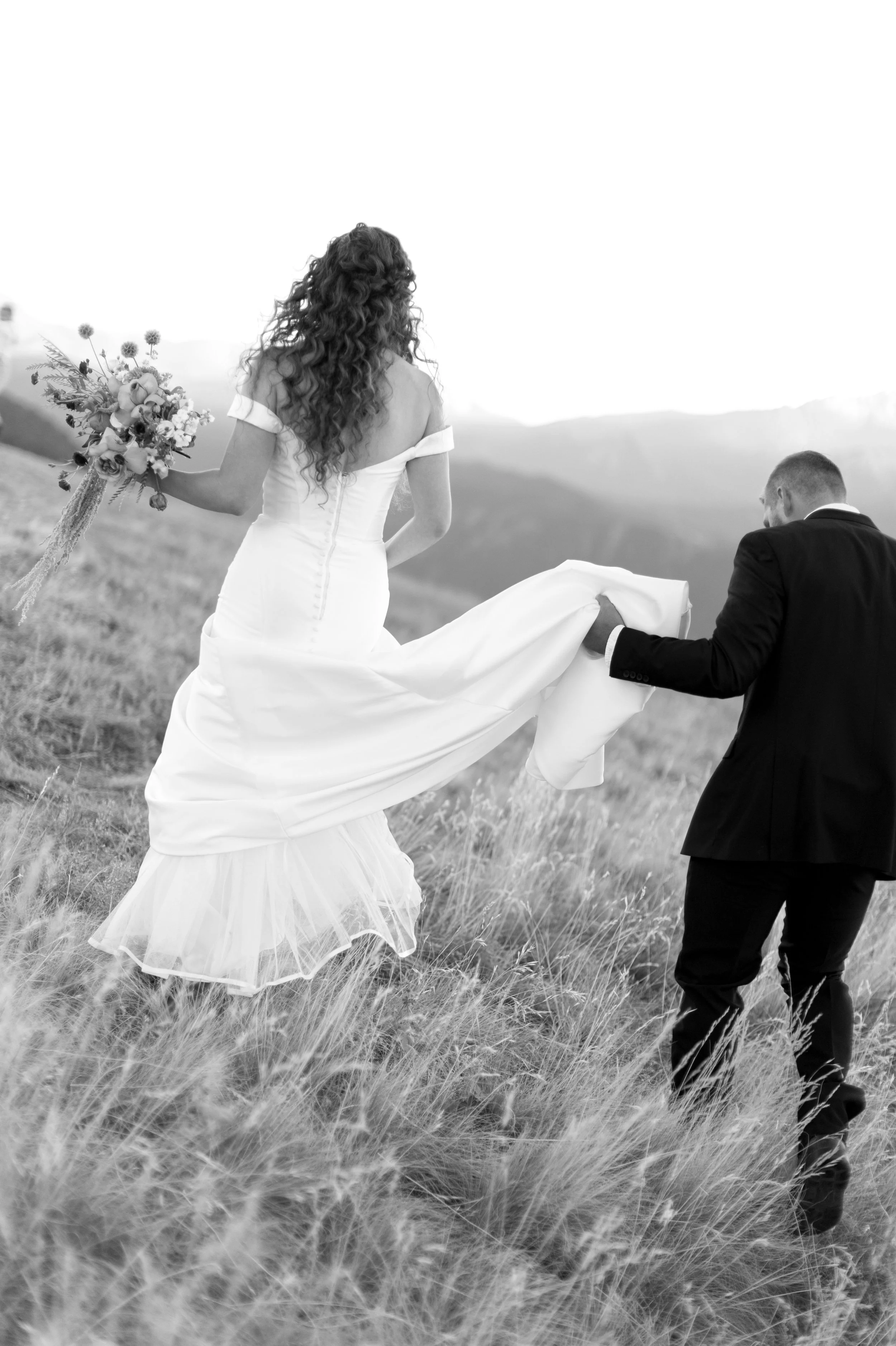 A bride wearing a wedding gown holding a bouquet of flowers, being carried by a groom in a suit, walking through a grassy field with mountains in the background.