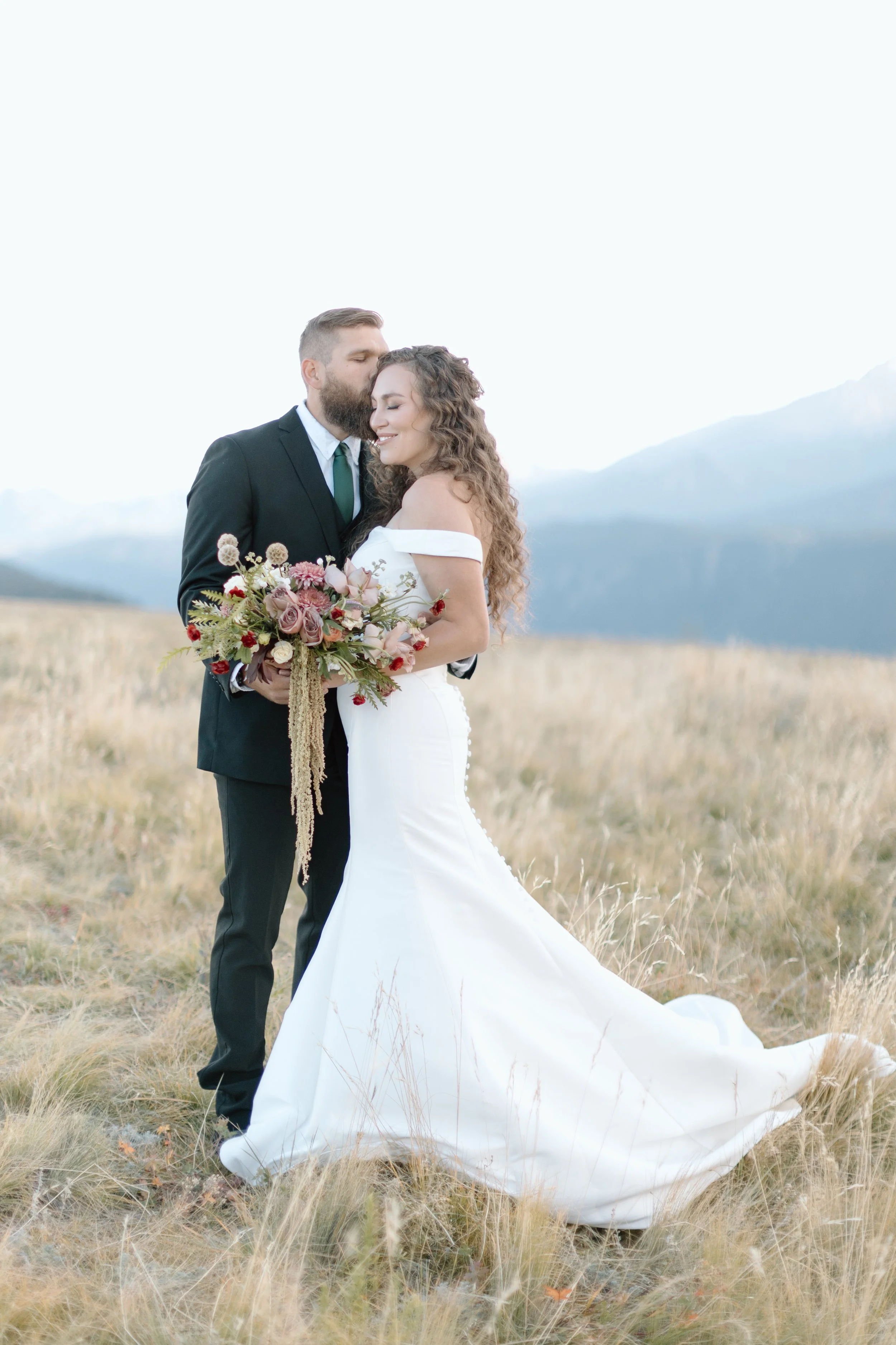 A bride and groom standing close together in a field with mountains in the background. The bride is in a white wedding dress holding a bouquet, and the groom is in a black suit with a green tie. The groom is kissing the bride on her temple, and they 